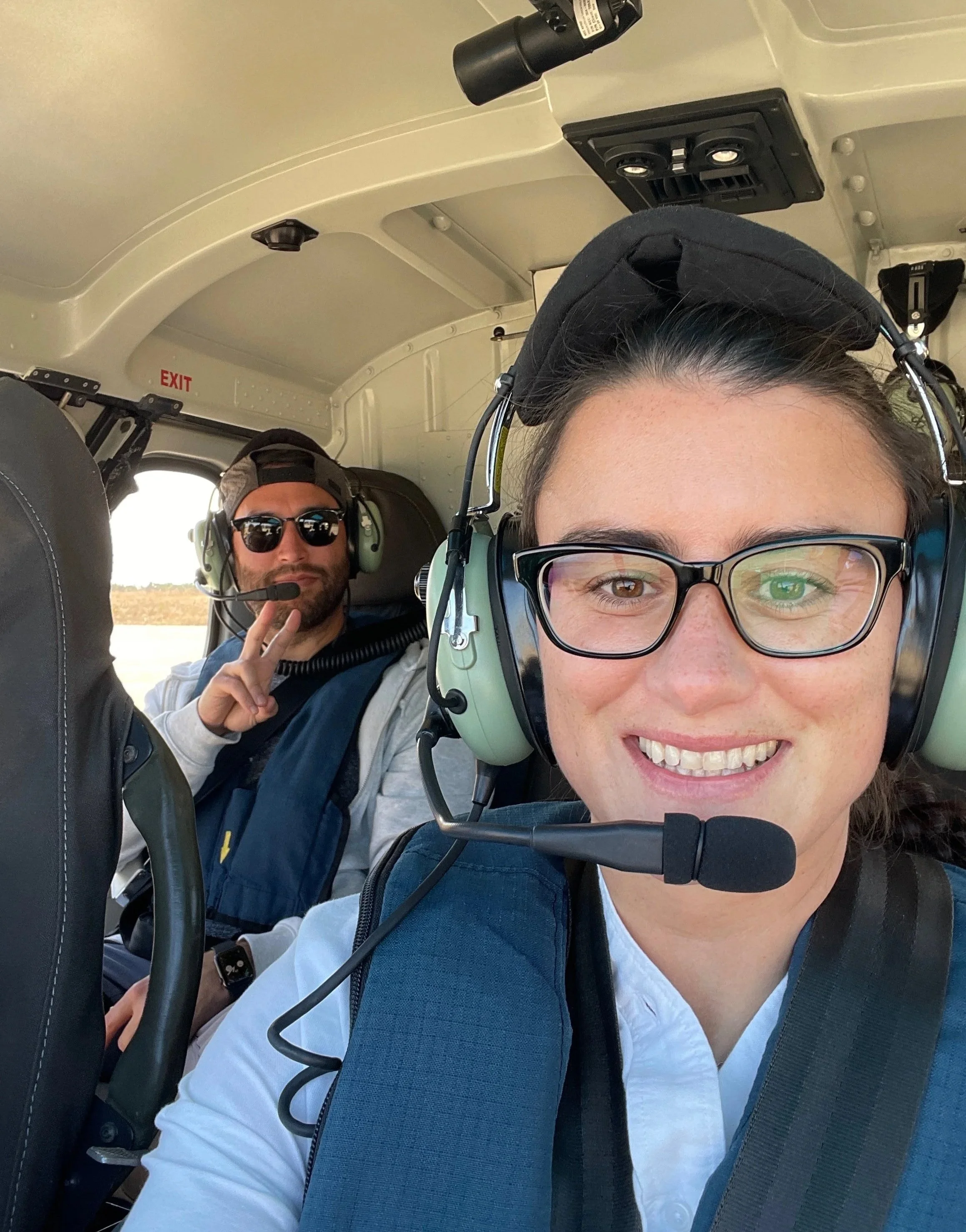 Two people inside a small aircraft cockpit, wearing headsets with microphones. The woman in the foreground has glasses, a white shirt, and is smiling. The man in the background is wearing sunglasses, a cap, and making a peace sign.