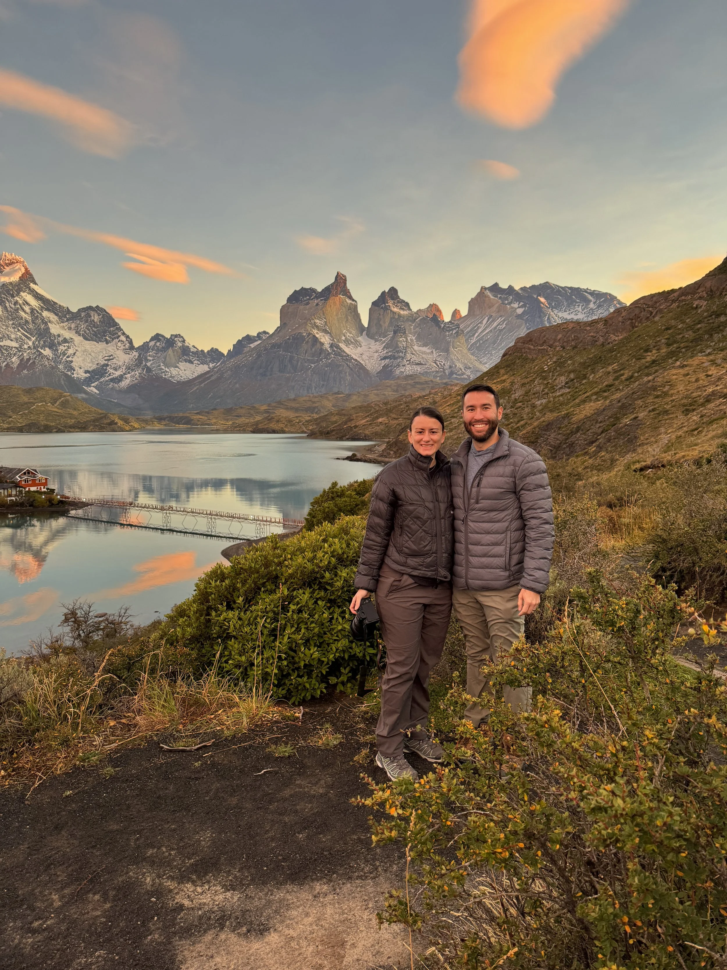 A smiling couple dressed in outdoor jackets standing together on a trail with a mountain lake and snow-capped mountains in the background during sunset.