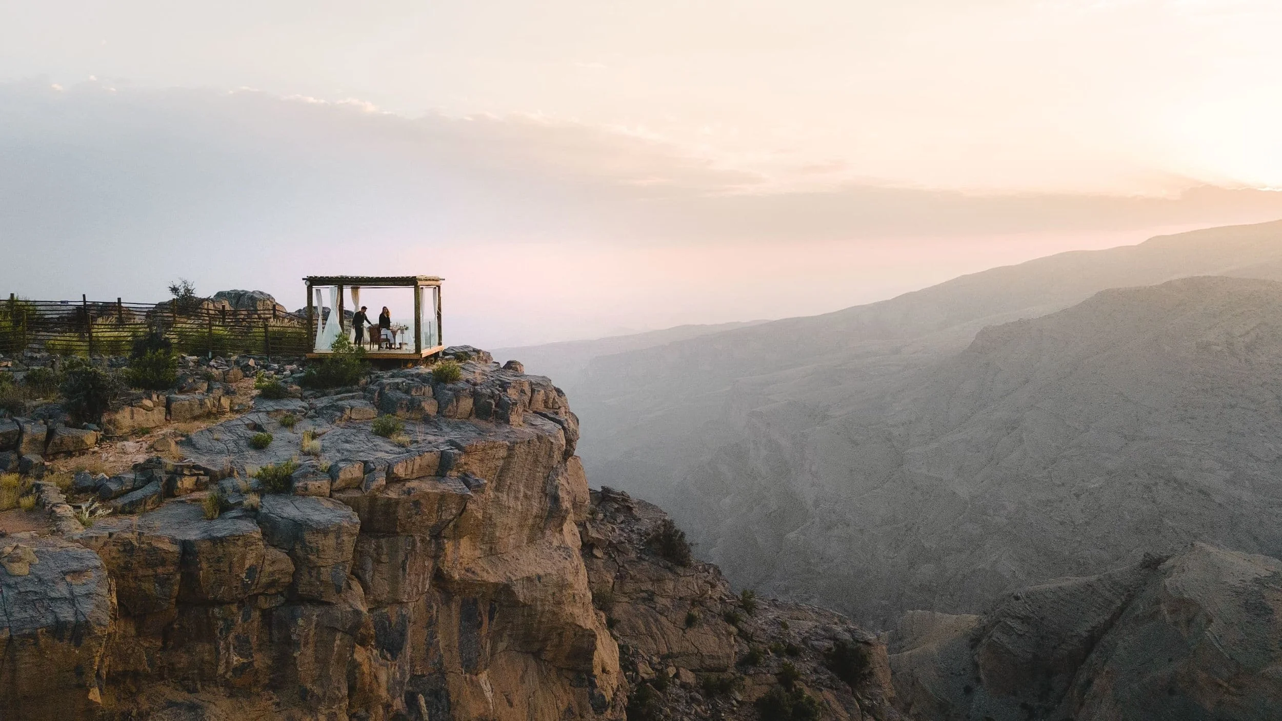 A small outdoor wooden patio with a canopy on a rocky cliff overlooking a vast canyon during sunset.