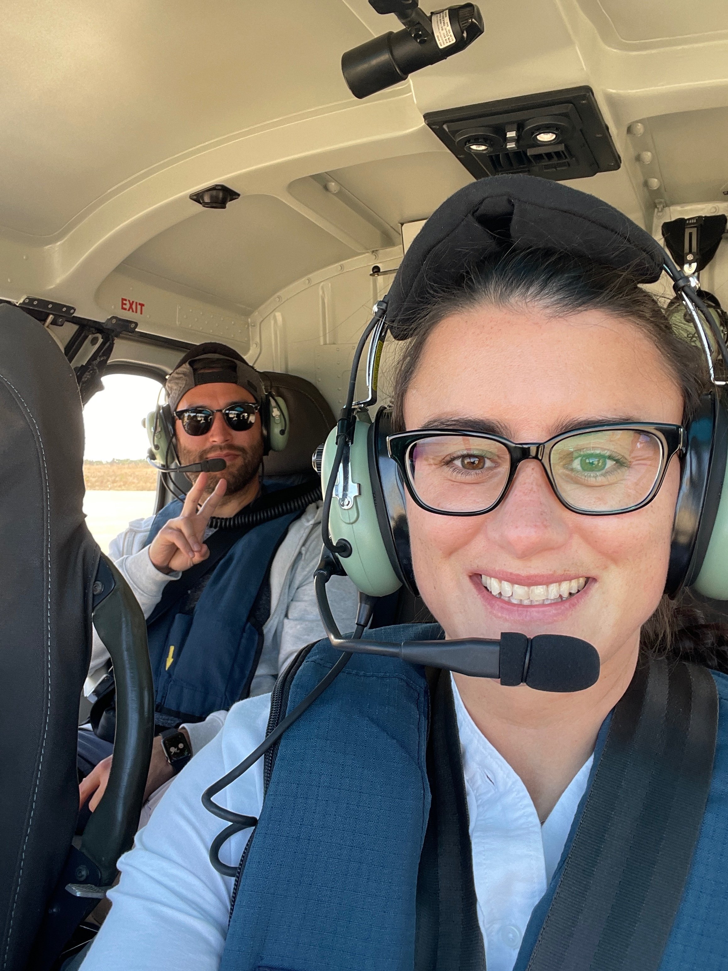 Two people inside a small aircraft cockpit, wearing headsets with microphones. The woman in the foreground has glasses, a white shirt, and is smiling. The man in the background is wearing sunglasses, a cap, and making a peace sign.