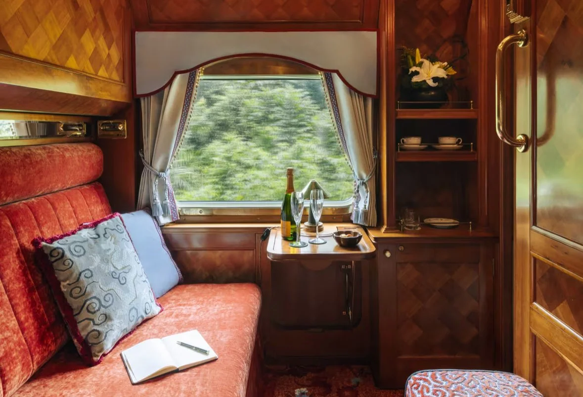 Inside a cozy train compartment with a window showing greenery outside. There is a red upholstered seat with decorative pillows, a small wooden table with champagne, glasses, and a bowl. Shelves contain cups and a flower arrangement.