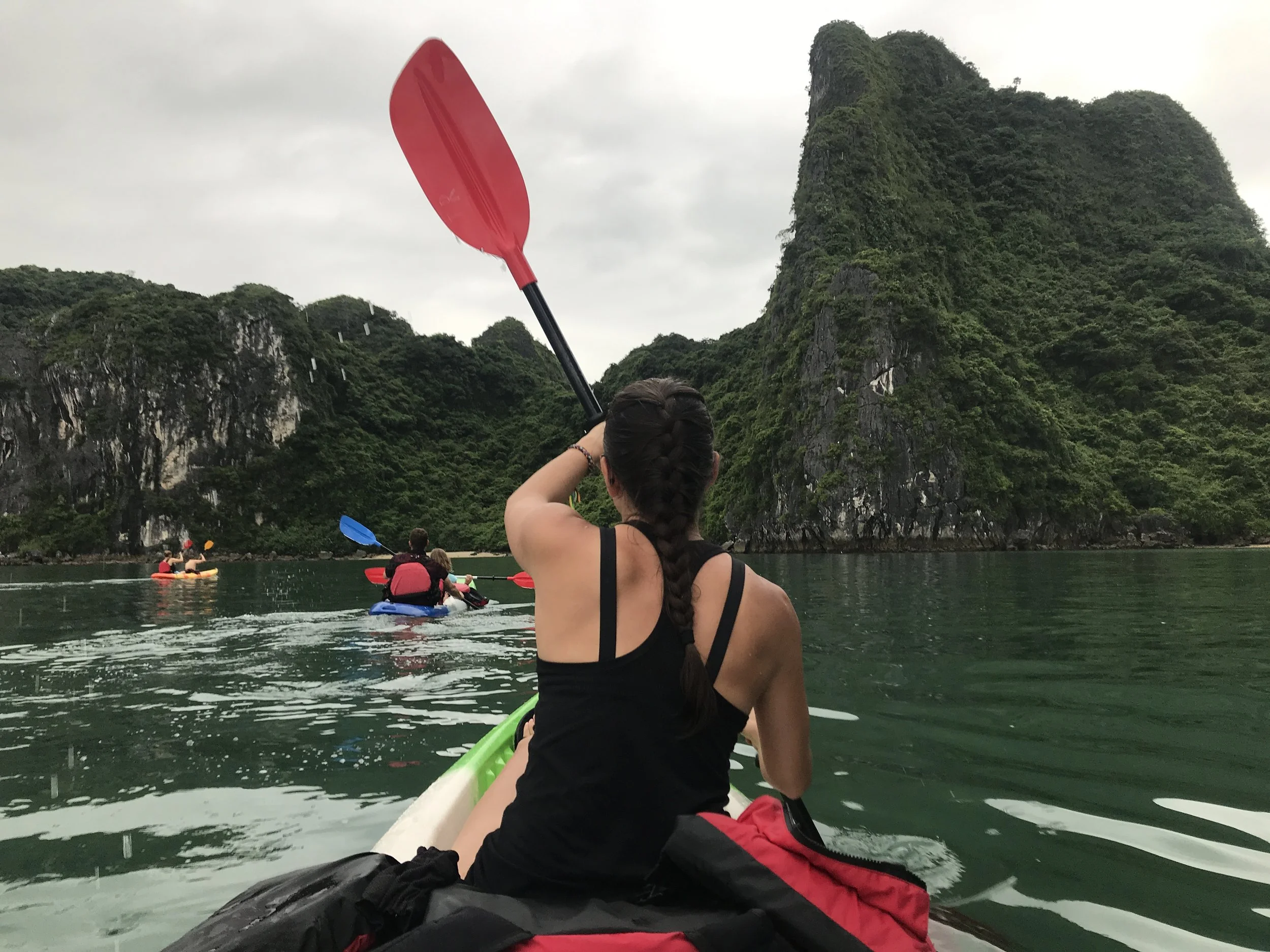 A woman with braided hair sitting in a kayak, paddling on a body of water surrounded by green mountainous cliffs, with other kayakers paddling in front of her.
