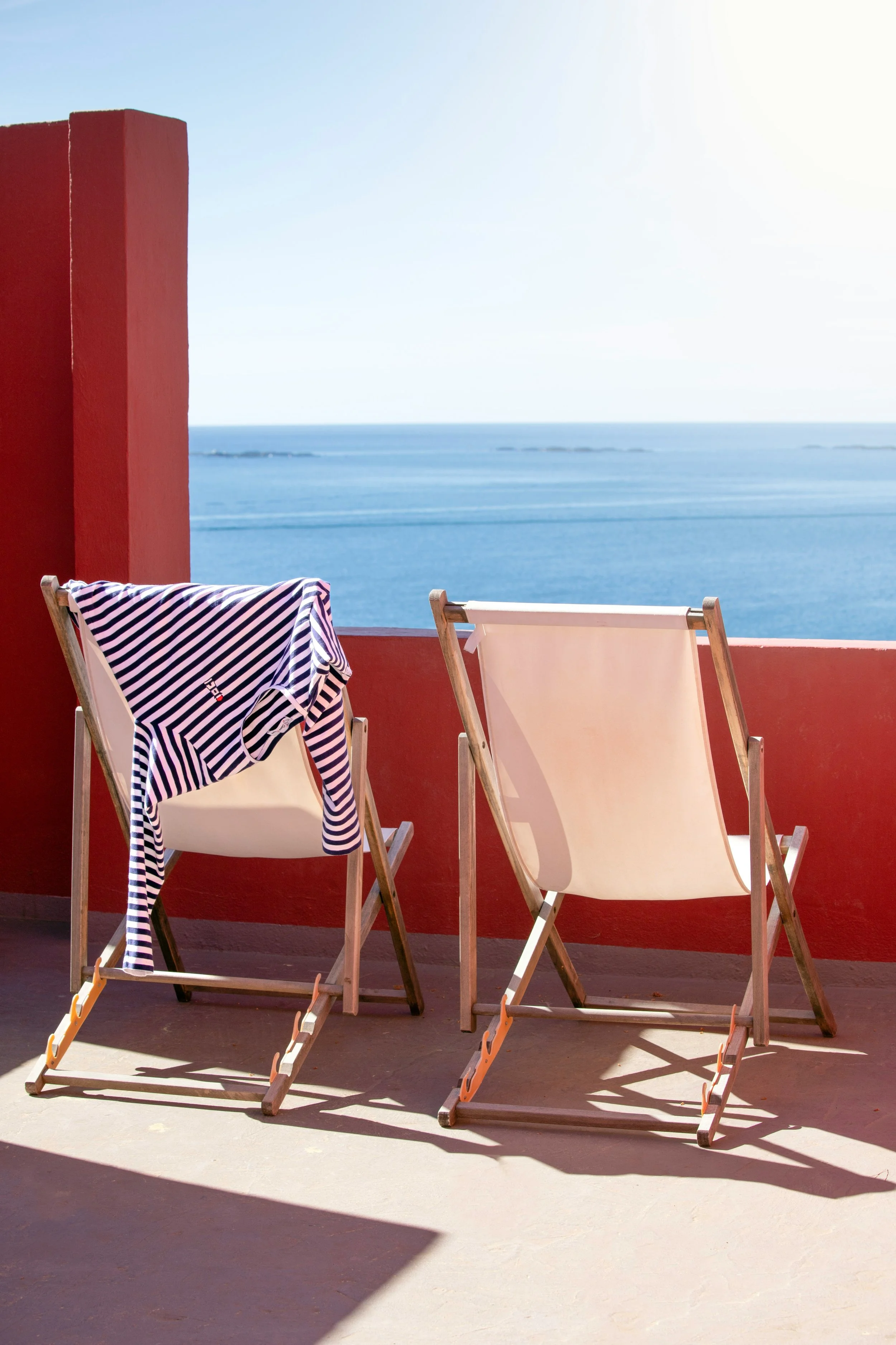 Two wooden deck chairs facing the ocean, one has a striped shirt draped over it, on a sunny balcony with red walls.