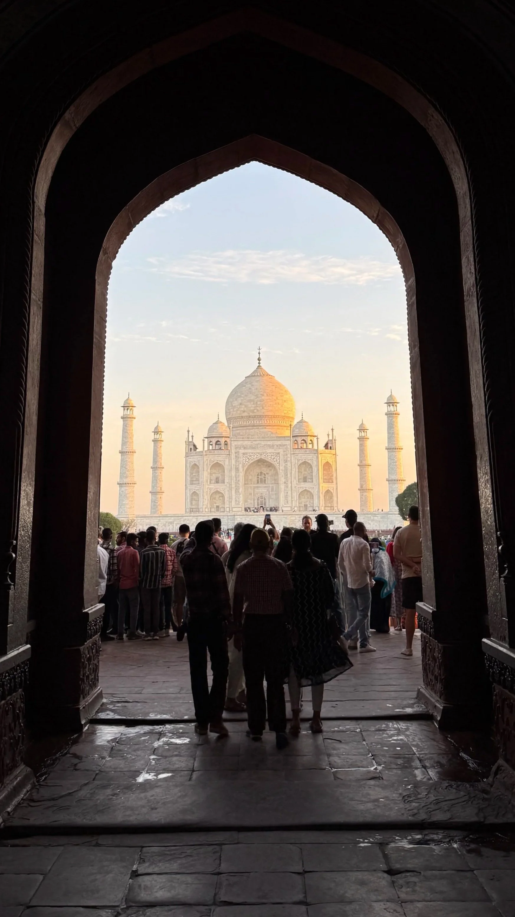 View of the Taj Mahal through an arch with tourists inside, capturing the iconic white marble mausoleum at sunset.