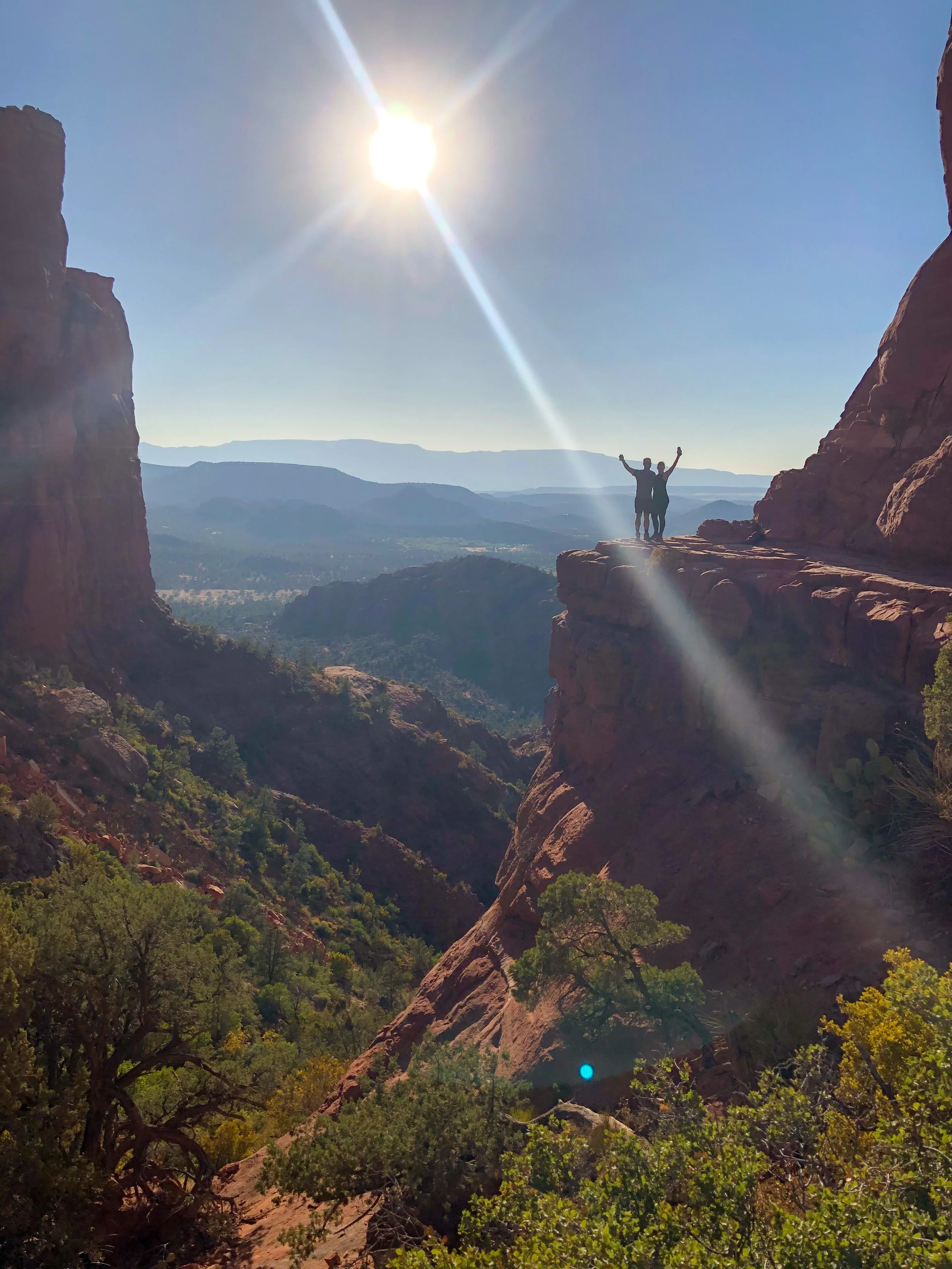 Two people standing on a rocky ledge in a canyon with mountains in the background and the sun shining brightly in the sky.