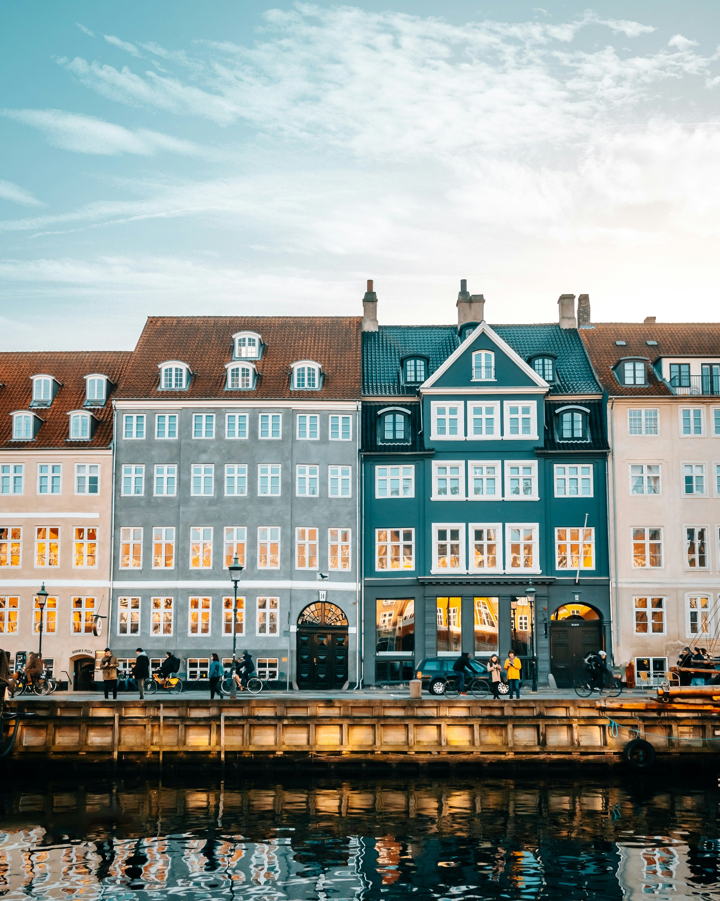 Colorful row of European-style buildings with large windows, situated by a waterway with reflections, under a partly cloudy sky.