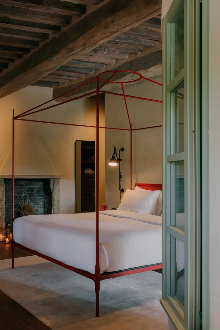 A bedroom with a red metal canopy bed, white bedding, and a wooden ceiling. There is a stone fireplace, a tall black cabinet, and a wall-mounted black reading lamp near the bed. The room has a cozy, rustic, and modern decor.