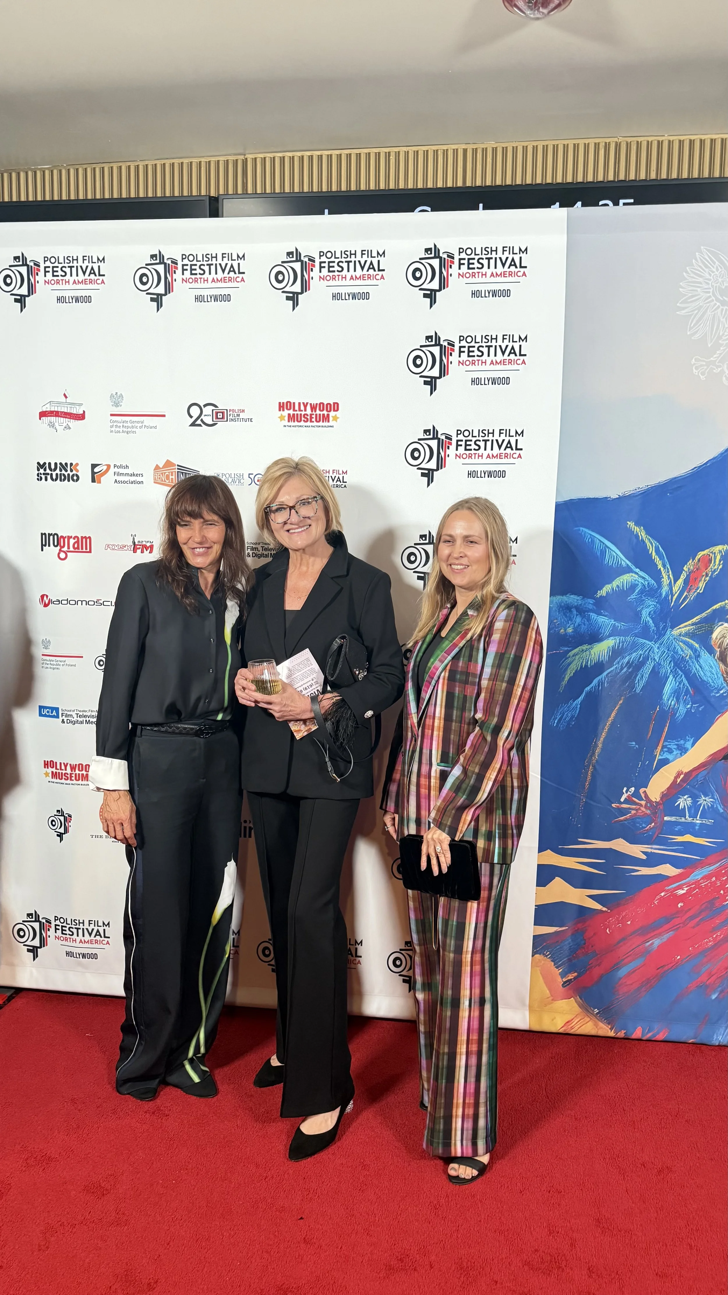Three women standing and smiling in front of a step and repeat backdrop at the Polish Film Festival North America event. One woman is holding a glass of wine, and the background features festival logos and a colorful poster. They are dressed in formal and colorful attire.
