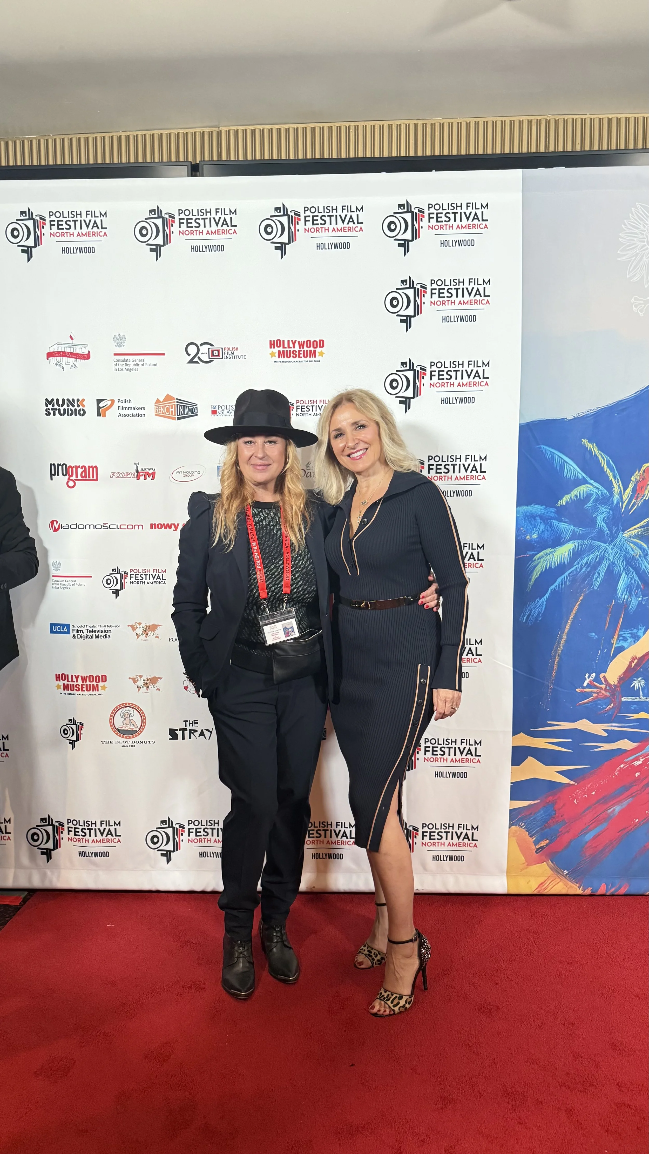 Two women standing together at the Polish Film Festival North America in Hollywood, posing for a photo on a red carpet in front of a step and repeat banner featuring the festival's logo and sponsors. One woman is wearing a black outfit with a hat, and the other is in a black dress with leopard print heels.