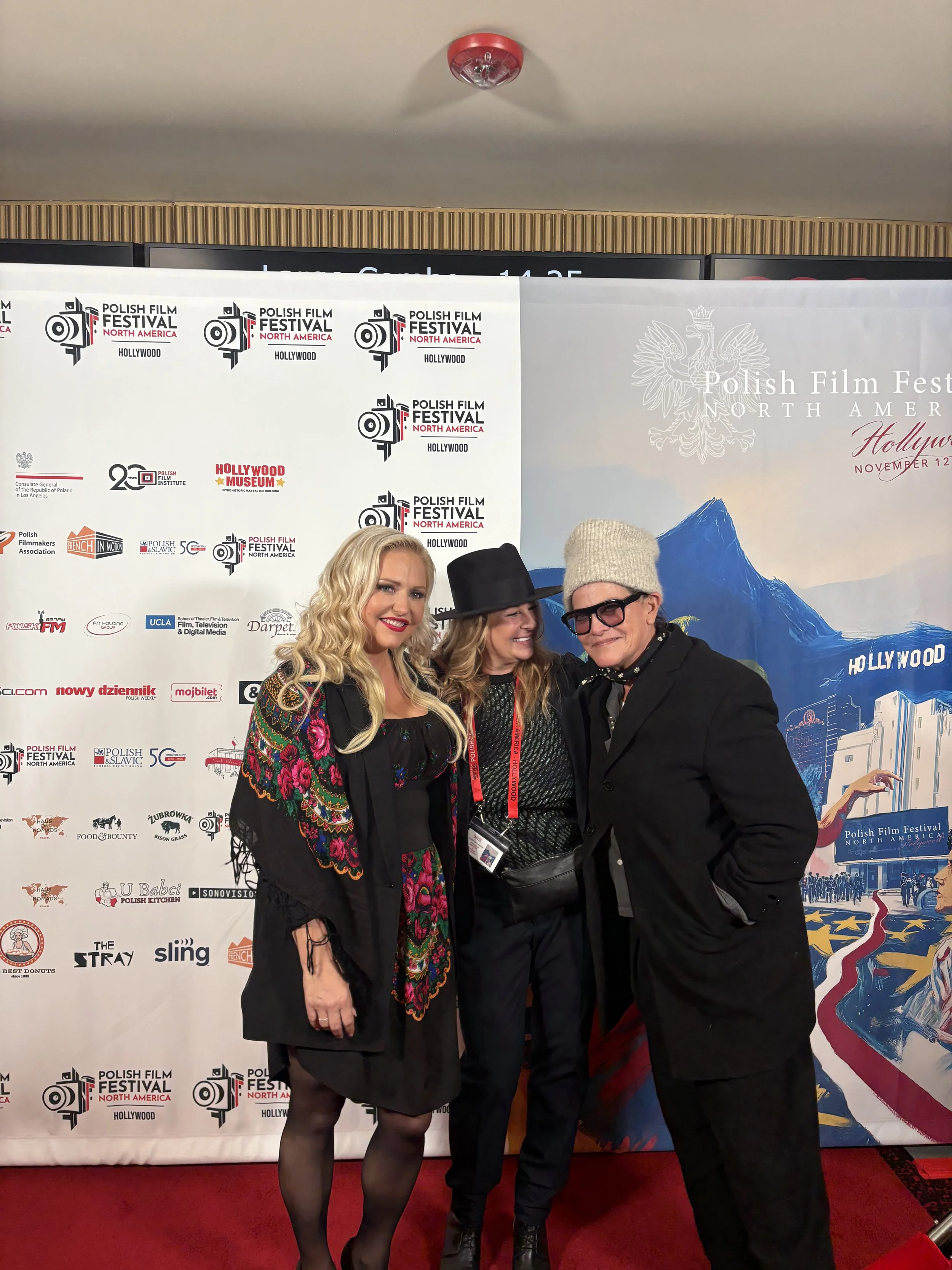 Three people standing on a red carpet at the Polish Film Festival North America in Hollywood. They are smiling and posing for a photo in front of a backdrop with various sponsor logos and festival branding.