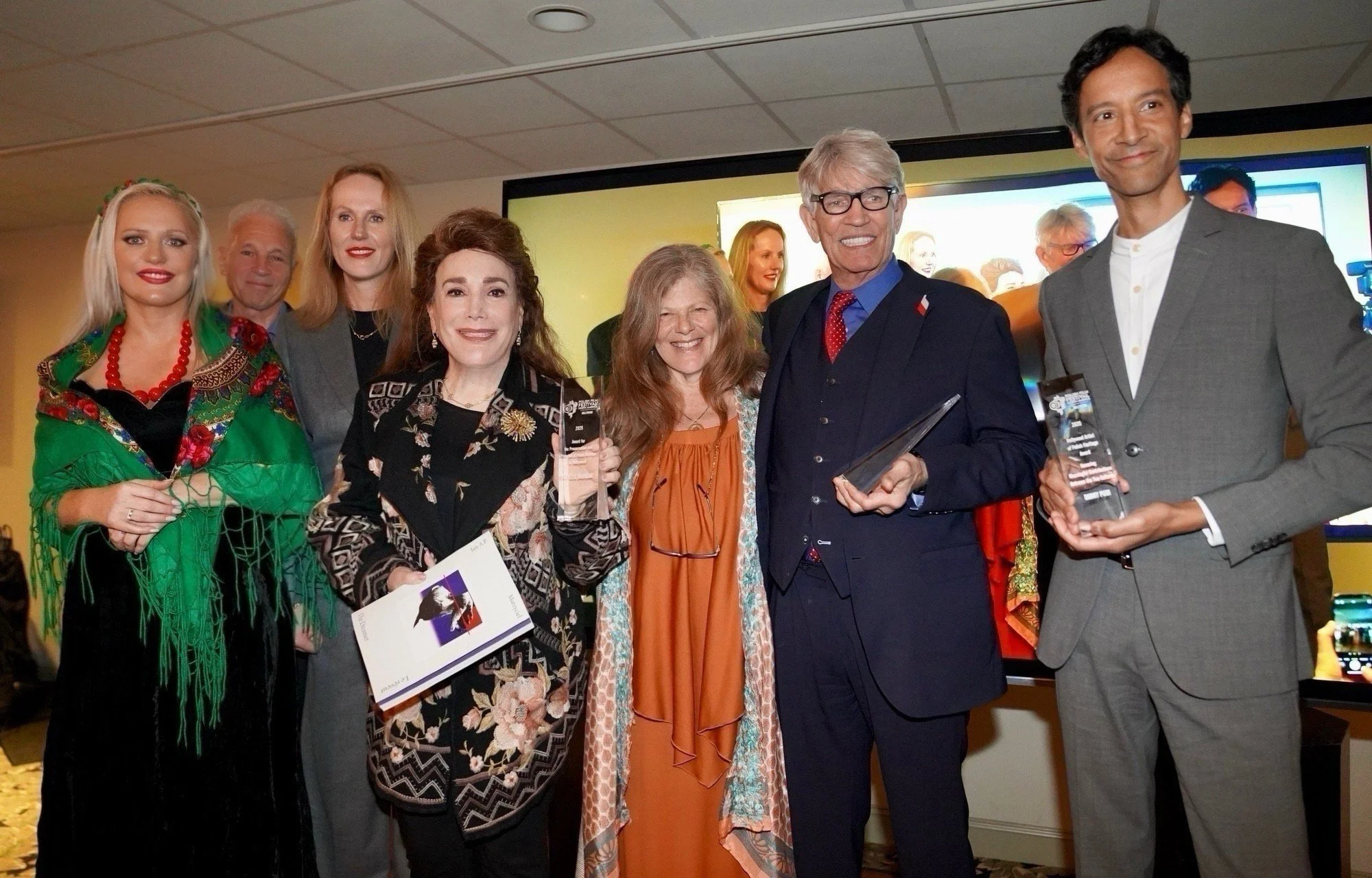 A group of seven diverse people standing together at an indoor event, some holding awards or certificates. They are smiling and dressed in a mix of formal and colorful attire.