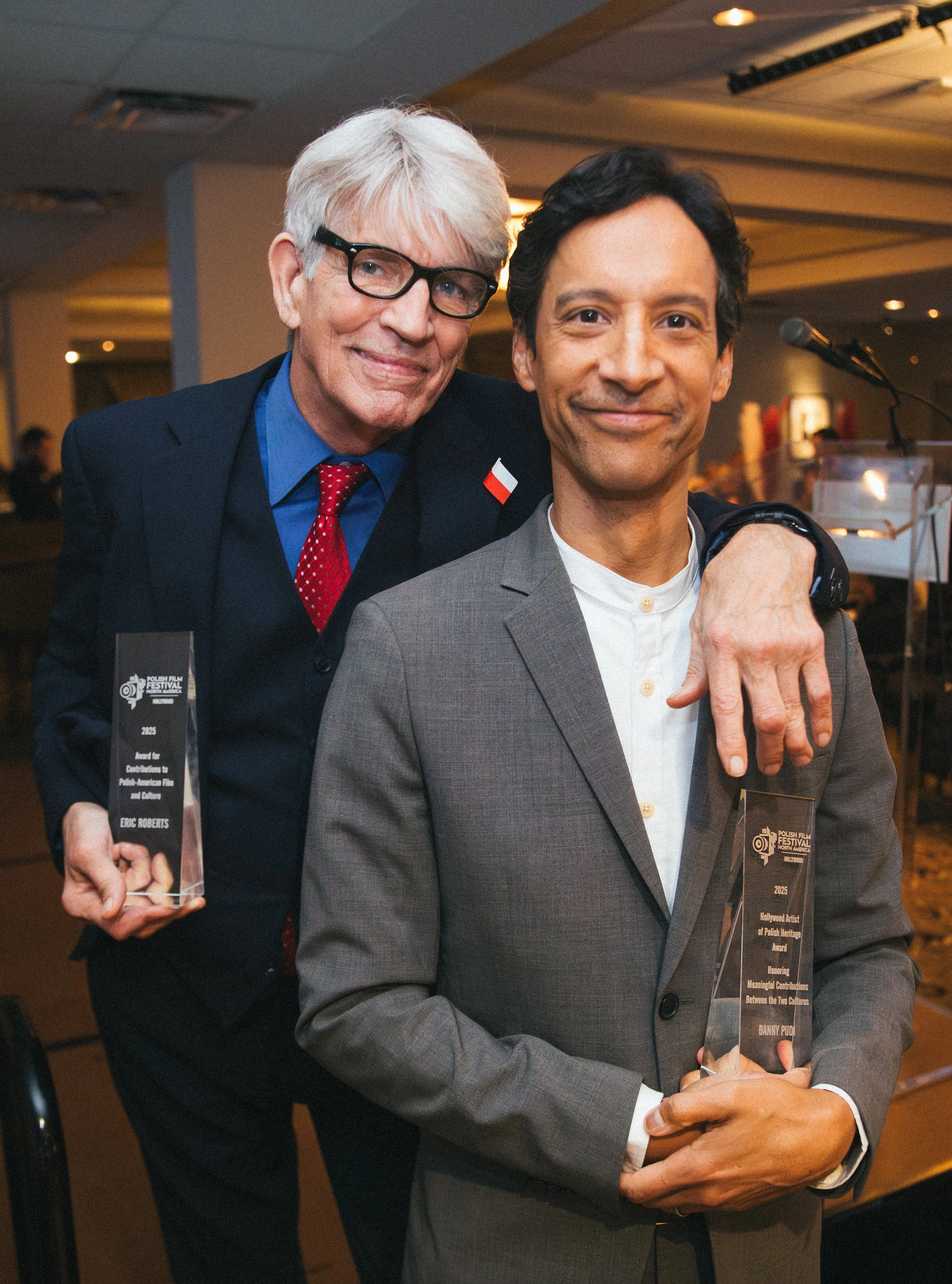 Two men at an awards event, one older with white hair and glasses, wearing a dark suit with a red tie, and the other younger with dark hair, wearing a gray suit, holding a glass award. The older man stands behind and to the side of the younger man, both smiling. The background shows a conference room with a podium and a microphone.