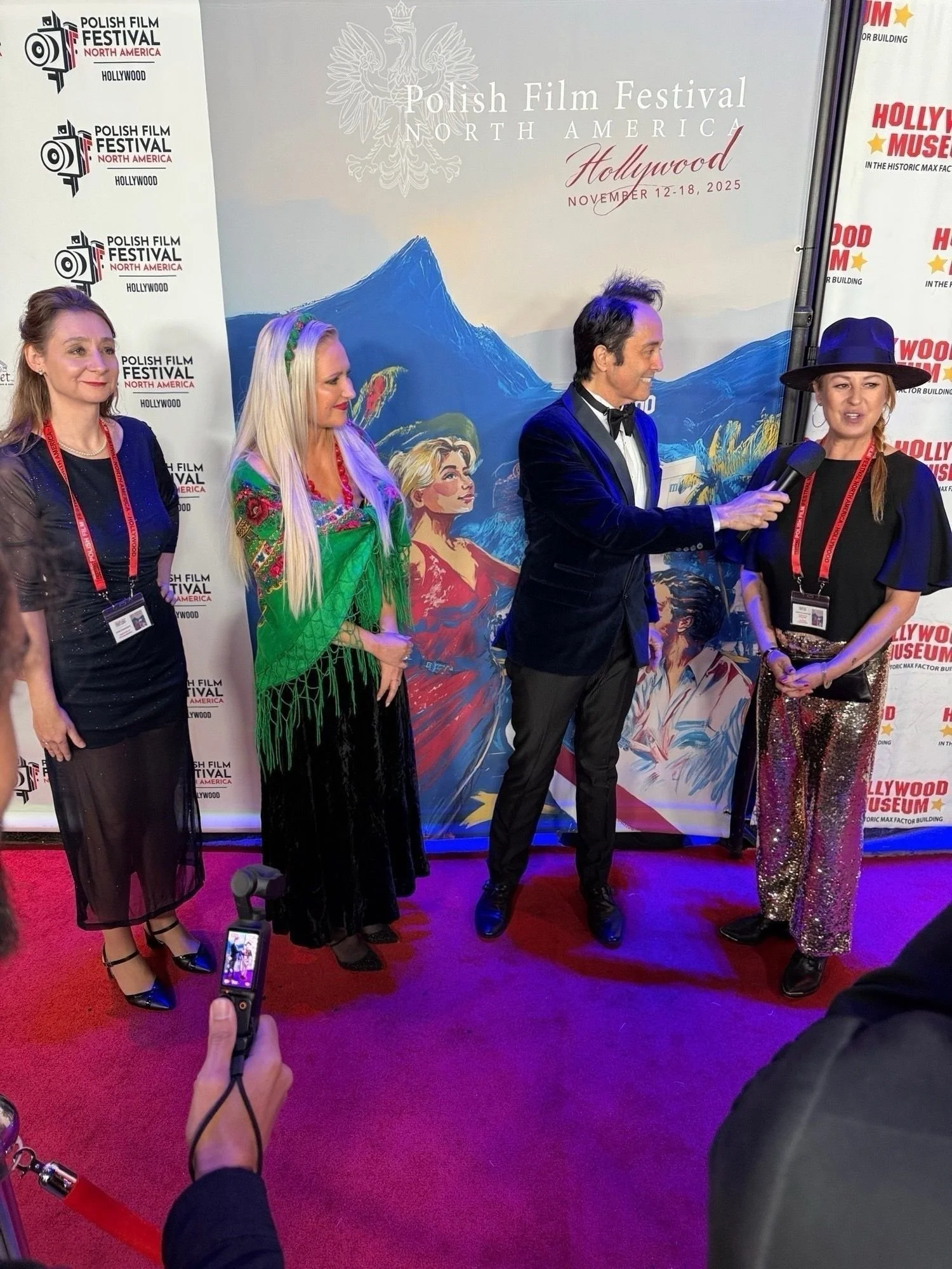 Four people standing on a red carpet at the Polish Film Festival North America in Hollywood, California. The background features promotional banners and artwork with mountains and a woman with blonde hair. A person in the foreground is taking a photo with a camera or phone.