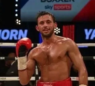 A shirtless male boxer in red boxing gloves and red shorts, standing in a boxing ring after a match, smiling with a victorious expression.