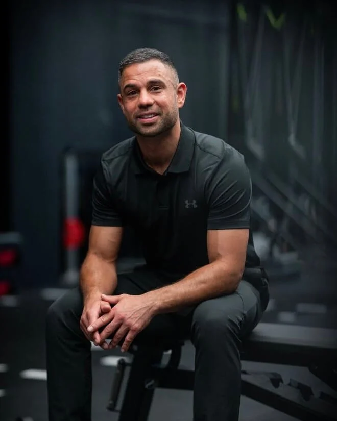 A man sitting on a workout bench in a gym, wearing a black Under Armour shirt and black pants, smiling at the camera.