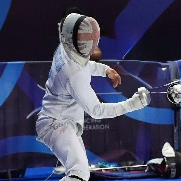 Fencer in white protective gear with a helmet featuring the Union Jack design, practicing or competing in a fencing match.