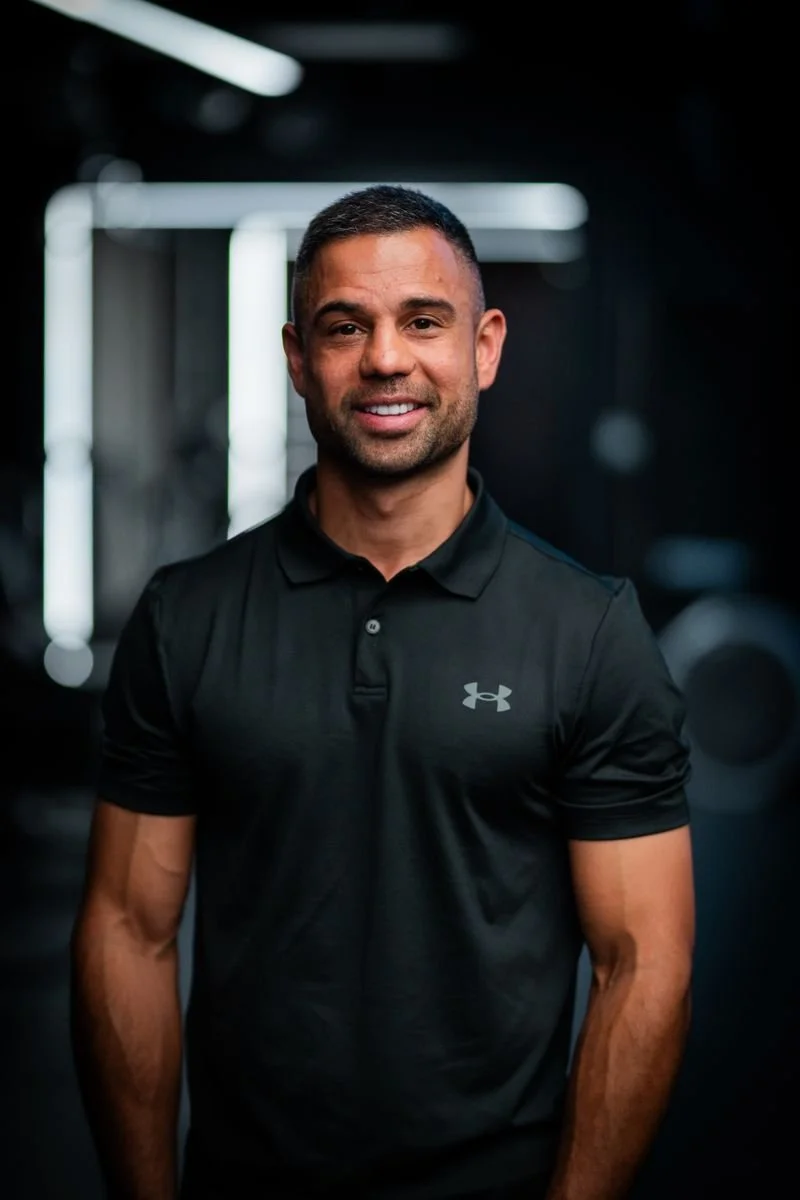 A man smiling, wearing a black Under Armour polo shirt, standing in a gym with blurred equipment and lighting in the background.