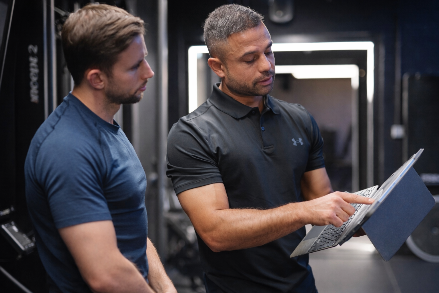 Two men in a gym, one with brown hair and a beard looking at a tablet, the other with short gray hair and a beard, wearing a black Under Armour shirt, pointing at the tablet.