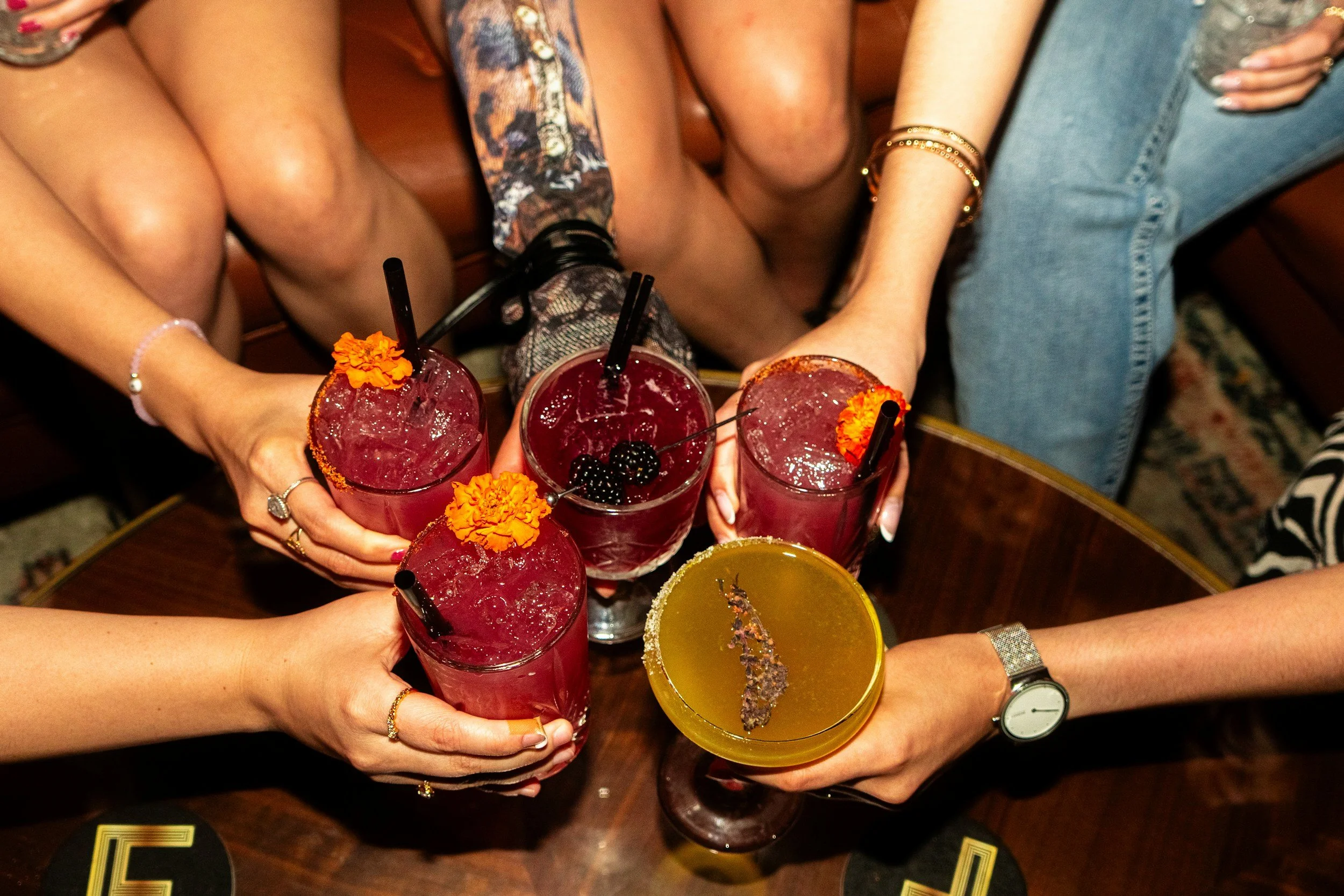 Four people raising glasses with cocktails at a wooden table in a bar or restaurant.
