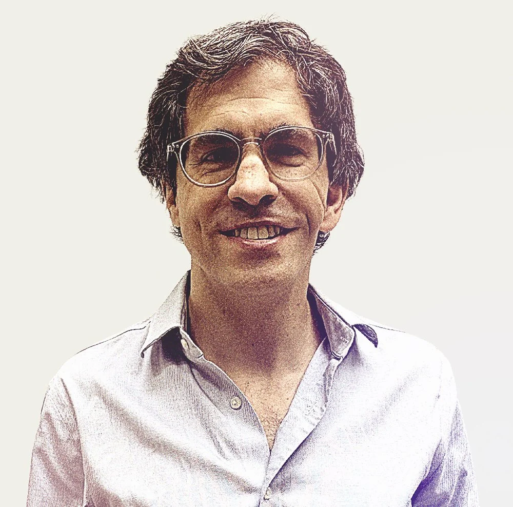 Hombre con gafas y cabello rizado, vistiendo una camisa de color claro y sonriendo.