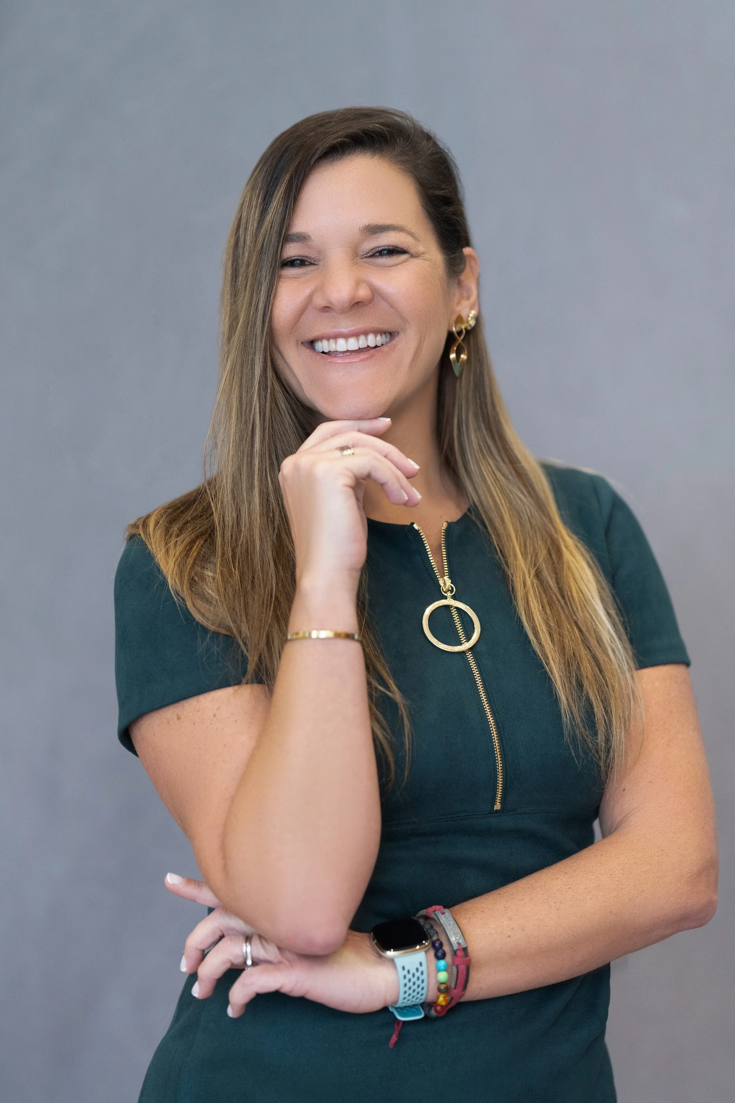 A woman with long brown hair smiling, wearing gold earrings, a green shirt with a zipper, and colorful bracelets, with arms crossed.