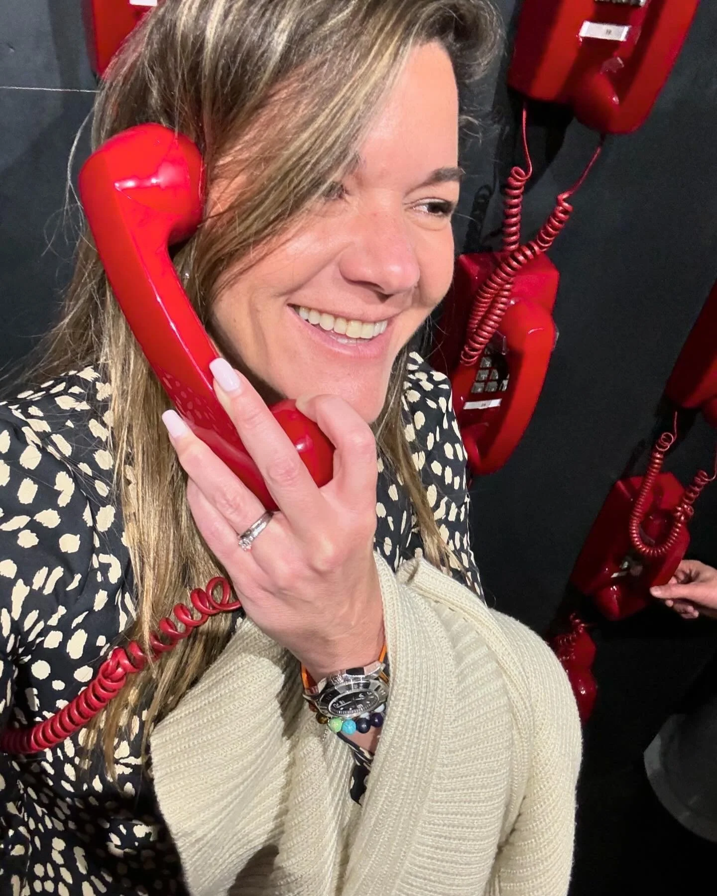 A woman with long, light brown hair is smiling while talking on a red rotary telephone. She wears a beige sweater, colorful beads, and a large watch. The background features red rotary telephones.