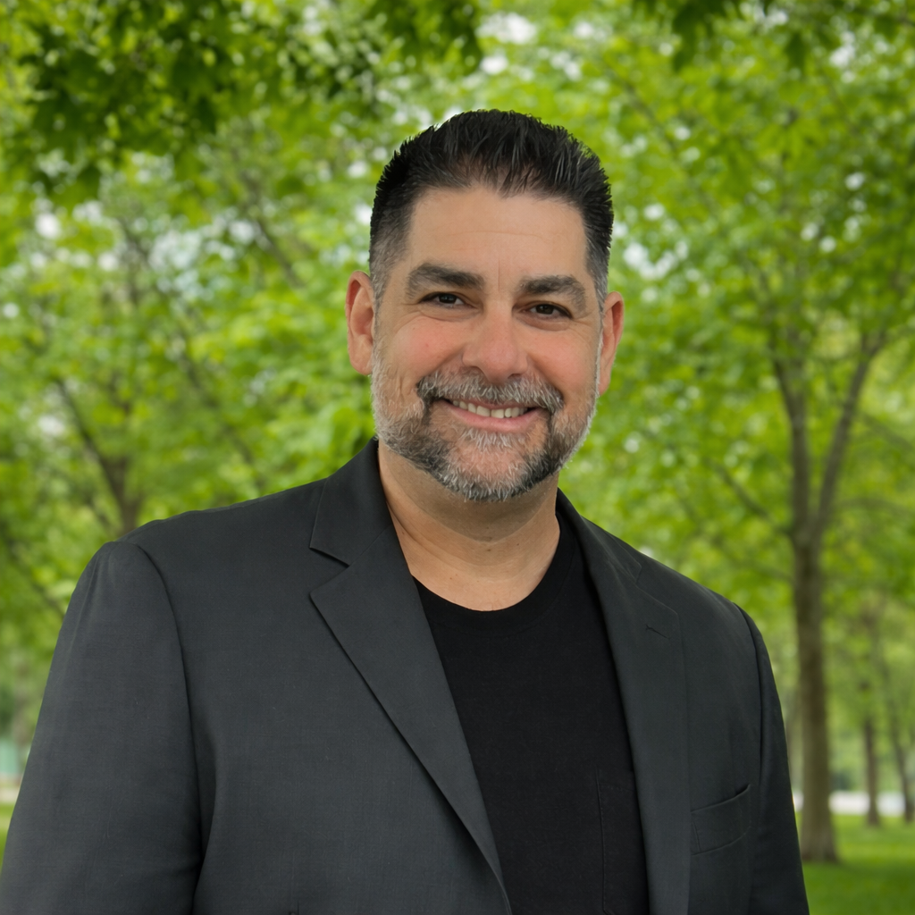 A man with short dark hair, a beard, and a mustache smiling in front of a background of green trees.