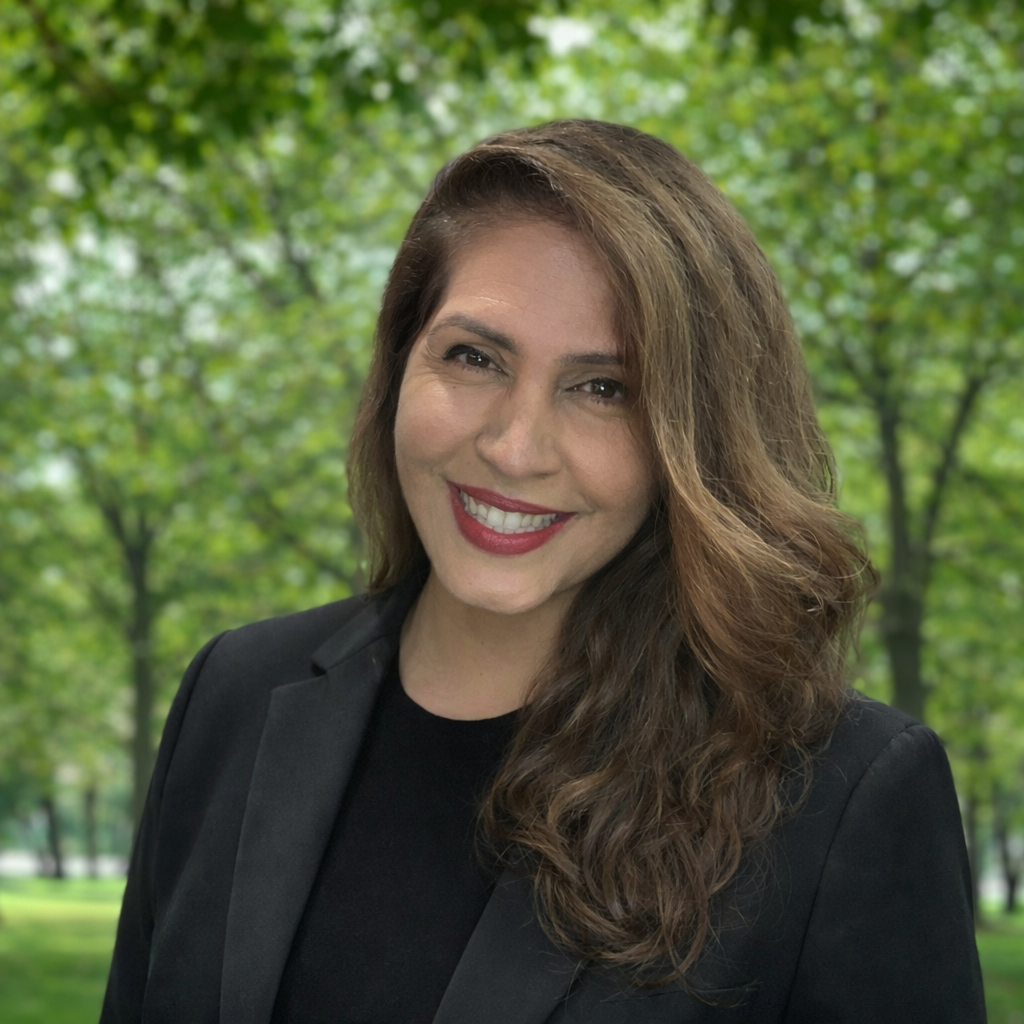 A woman smiling outdoors with green trees in the background.