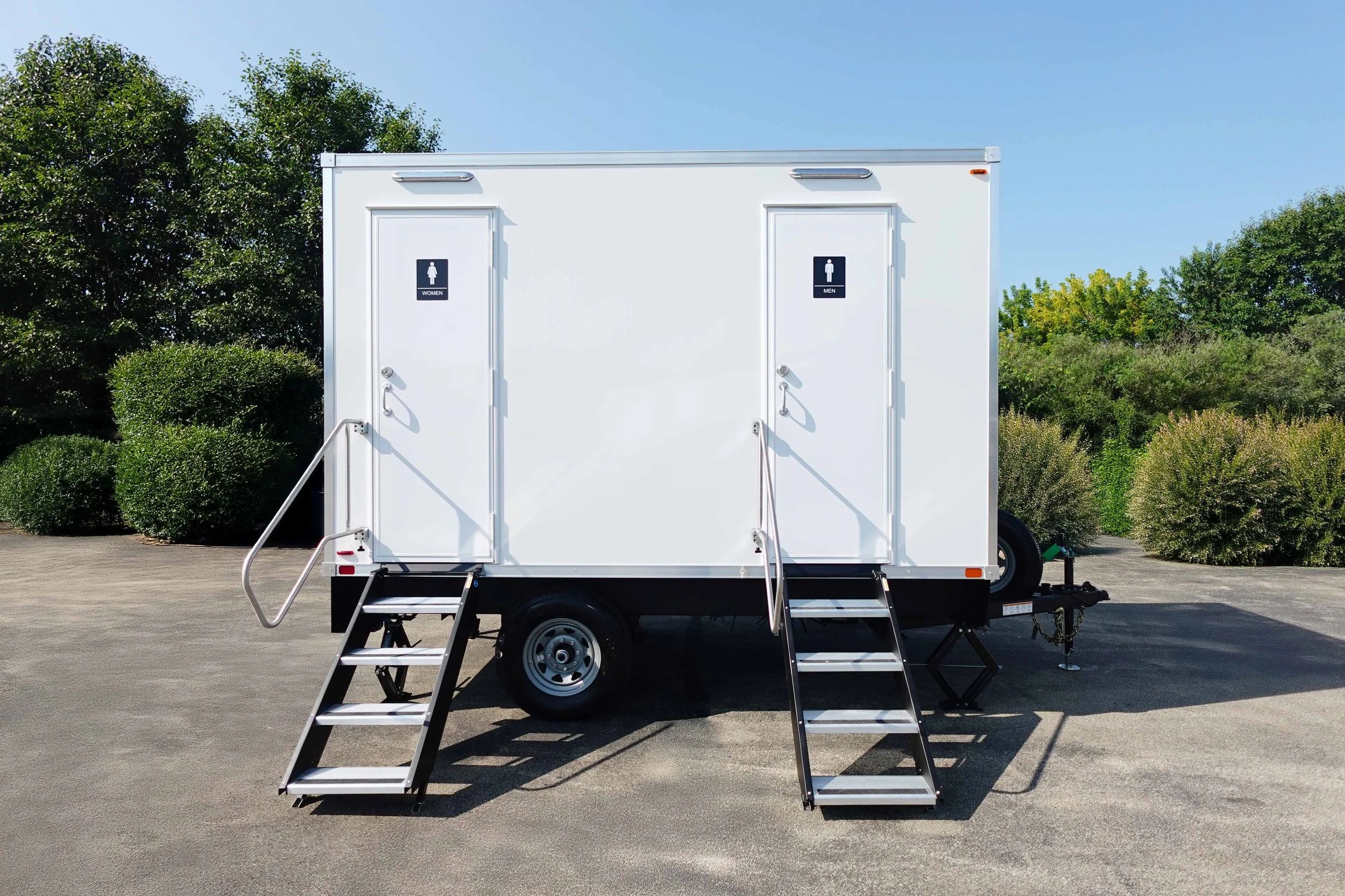 White mobile restroom trailer with two doors marked for women and men, each with stairs and handrails, parked on pavement with greenery in the background.