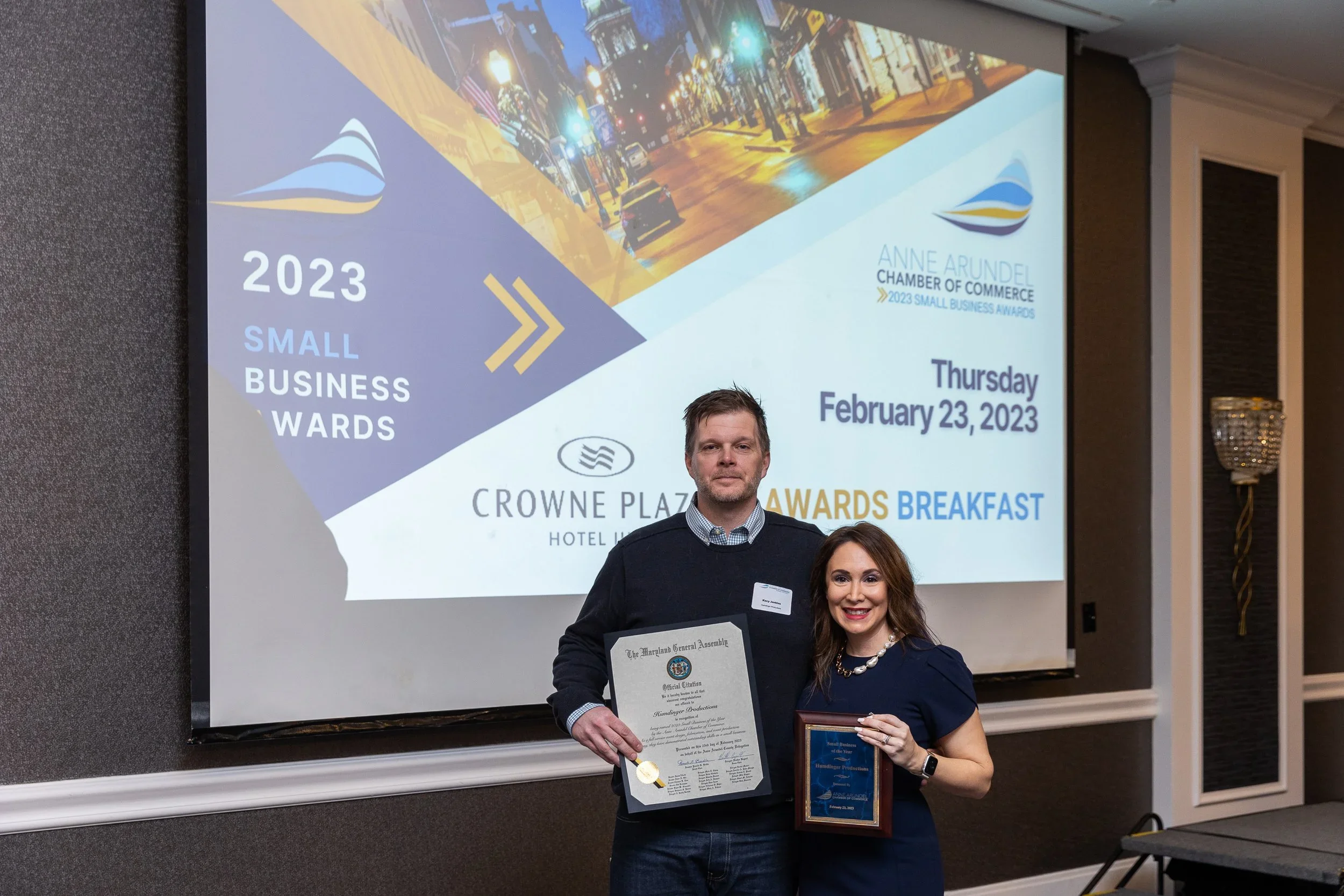 A man and woman standing together at an awards event in front of a large screen. The man is holding a certificate, and the woman is holding a plaque. The screen behind them shows details of the event, including the date February 23, 2023, and mention