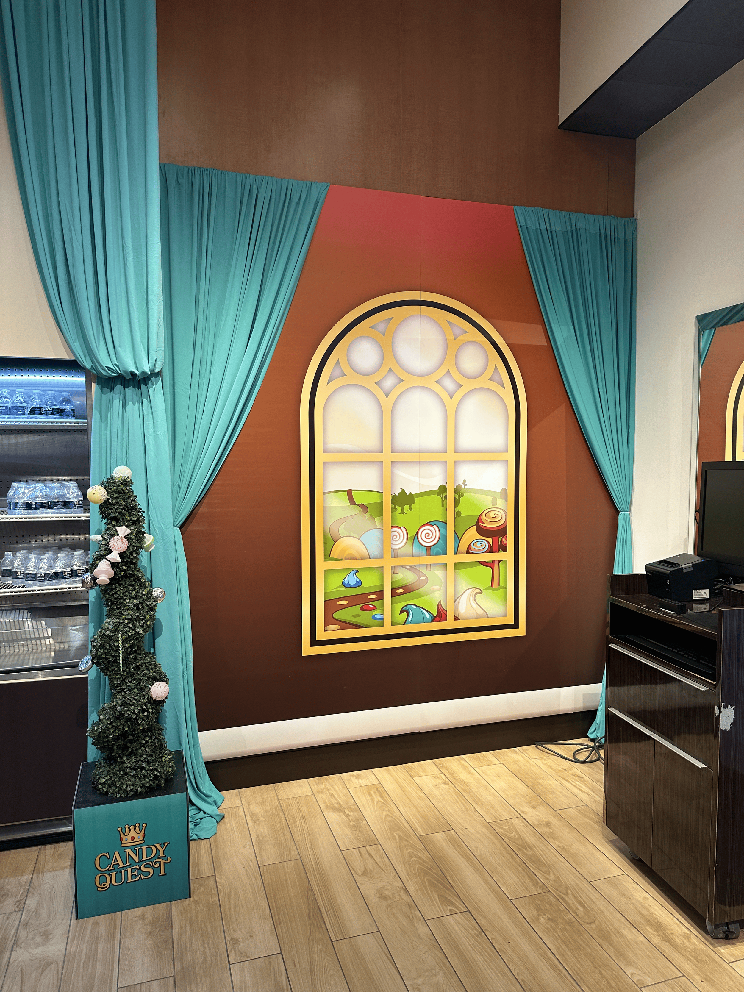 Decorative scene with teal curtains framing a colorful window mural of a fantasy landscape, and a small topiary bush decorated with ornaments labeled 'Candy Quest' on a sign.