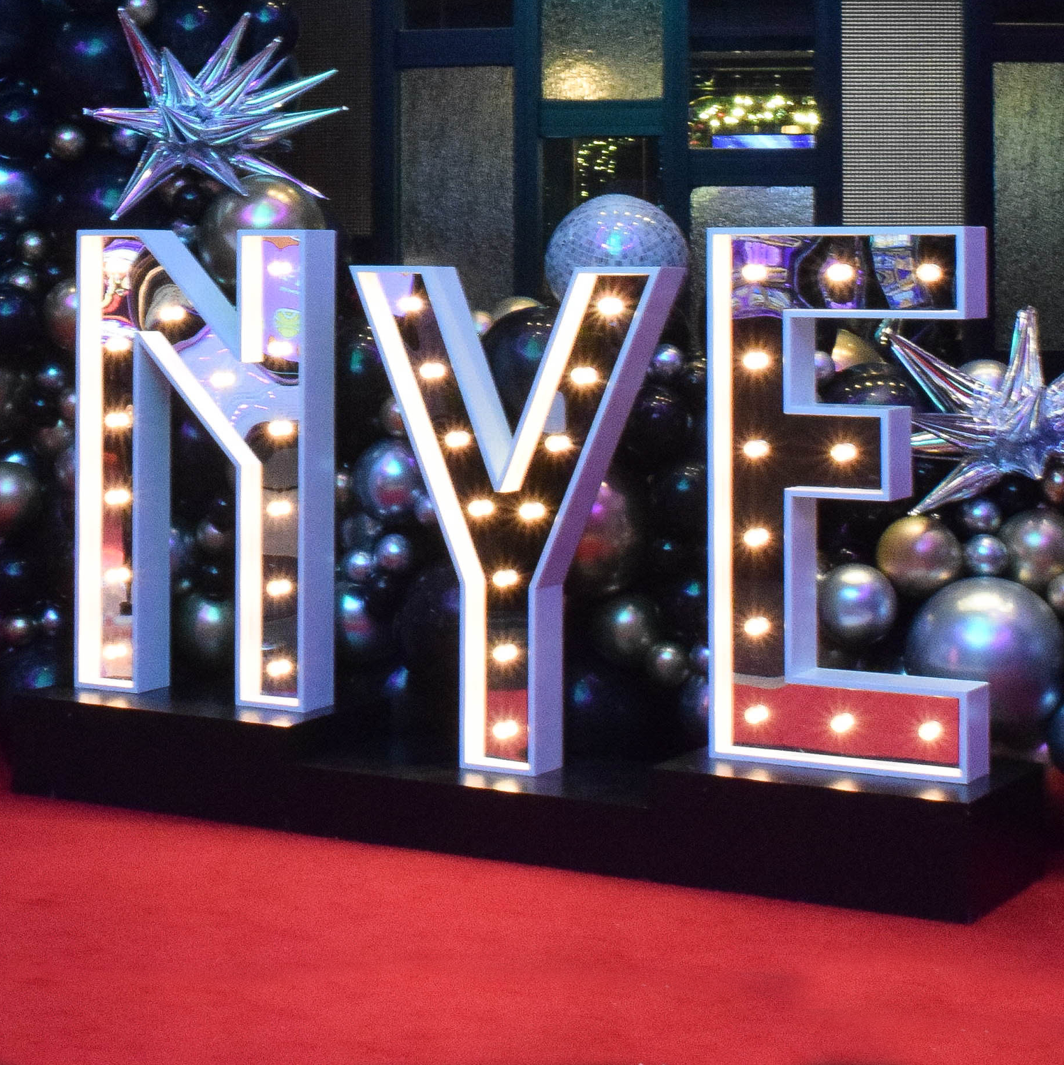 Decorative display spelling out the word "LOVE" with large illuminated letters, surrounded by silver, gold, and black Christmas ornaments, and large star-shaped decorations, on a red table at a festive celebration.