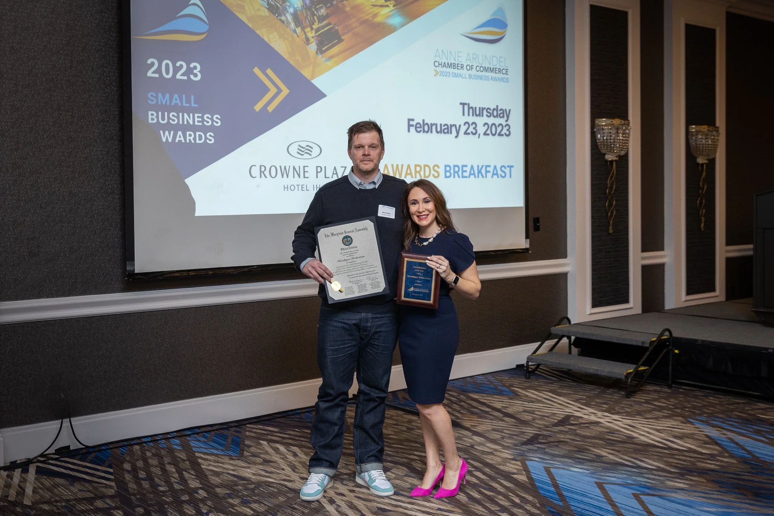 A man and a woman standing together on a stage, holding awards, during a banquet event in a hotel conference room. In the background is a large projection screen displaying details about the 2023 Small Business Awards breakfast, held on February 23, 
