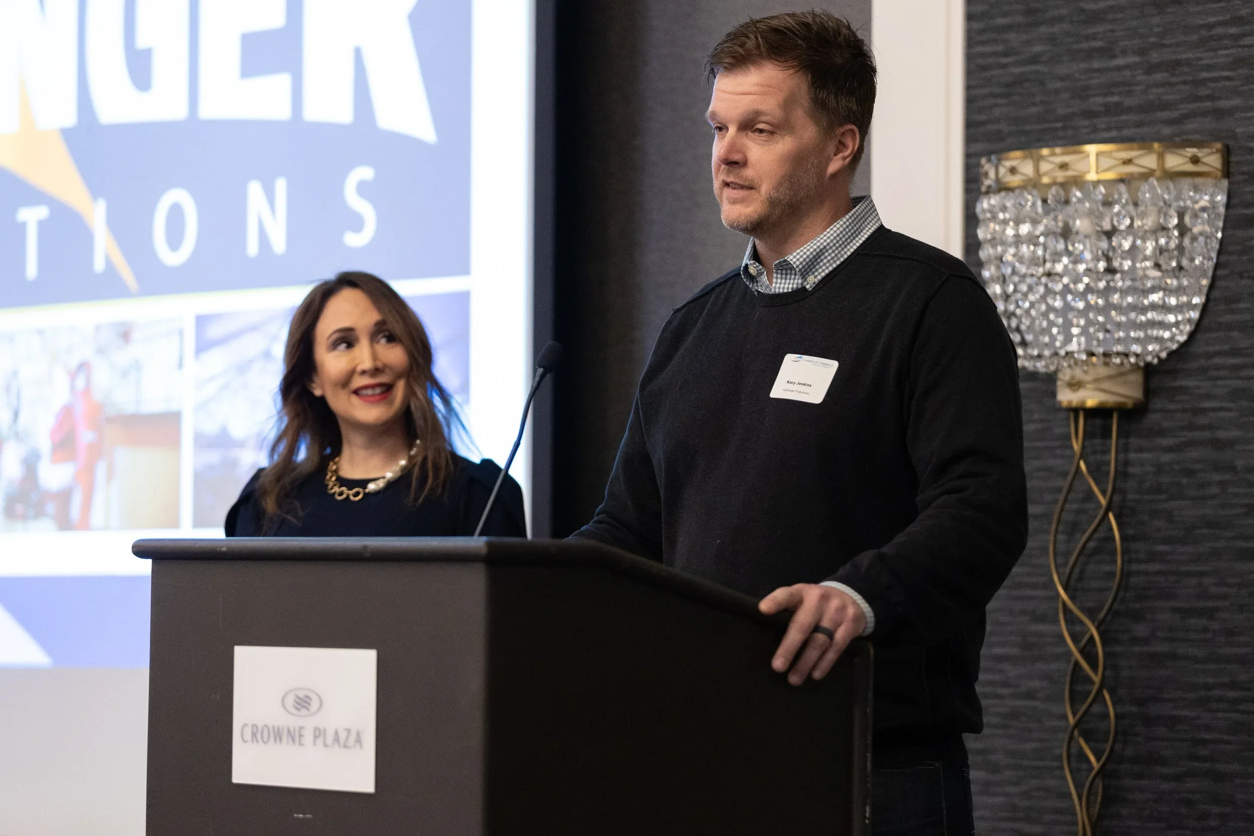 A woman and a man standing at a podium during a presentation at Crowne Plaza hotel. The woman is looking at the man with a smile, and the man is speaking into the microphone.