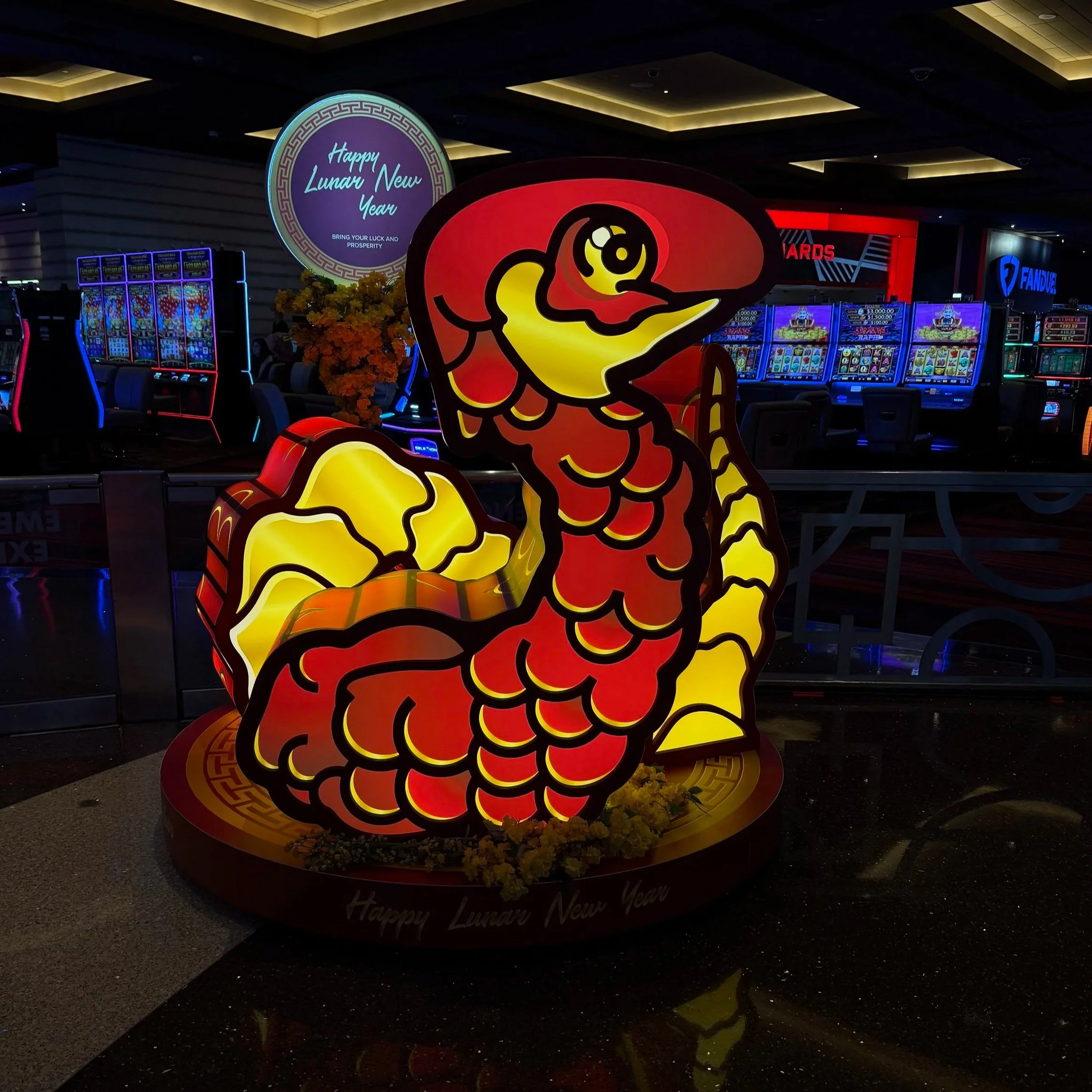 Illuminated decorative display of a cartoon-style red and yellow fish for Lunar New Year, with a sign wishing Happy Lunar New Year and prosperity, set inside a casino or gaming area with slot machines in the background.