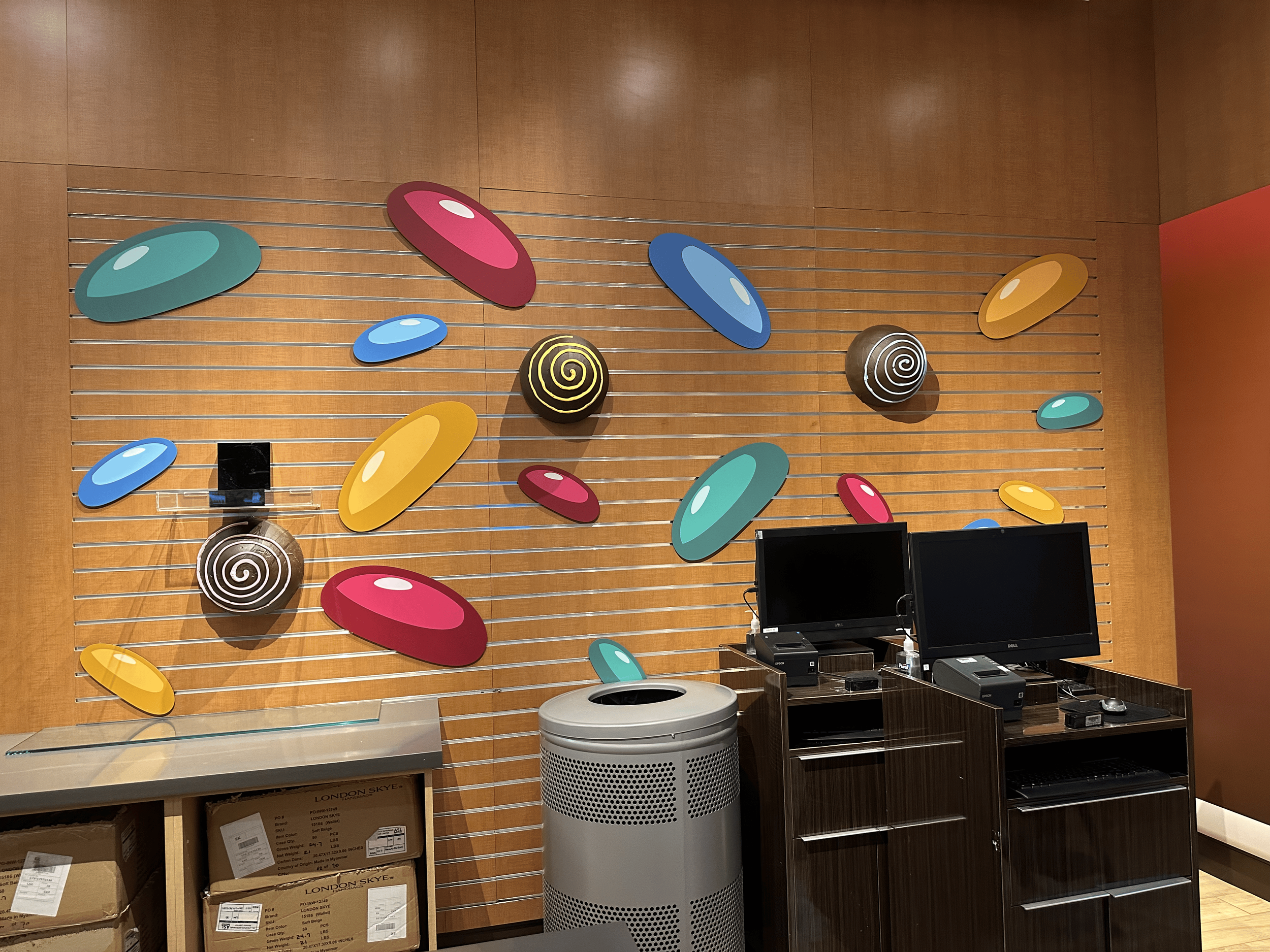 Decorative wall with colorful oval and spiral art pieces, a black wall-mounted camera, two computer monitors, a computer tower, and various electronic accessories.