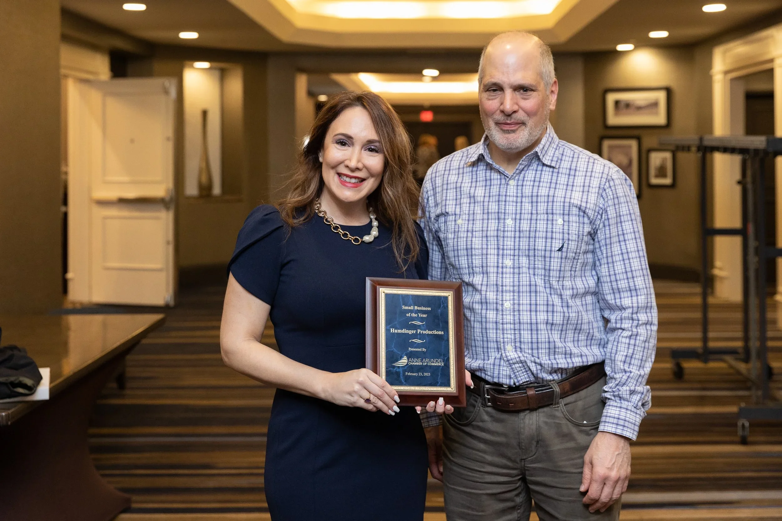A woman in a navy blue dress holding a framed award, standing beside a man in a plaid shirt in an indoor event space with framed pictures on the wall.
