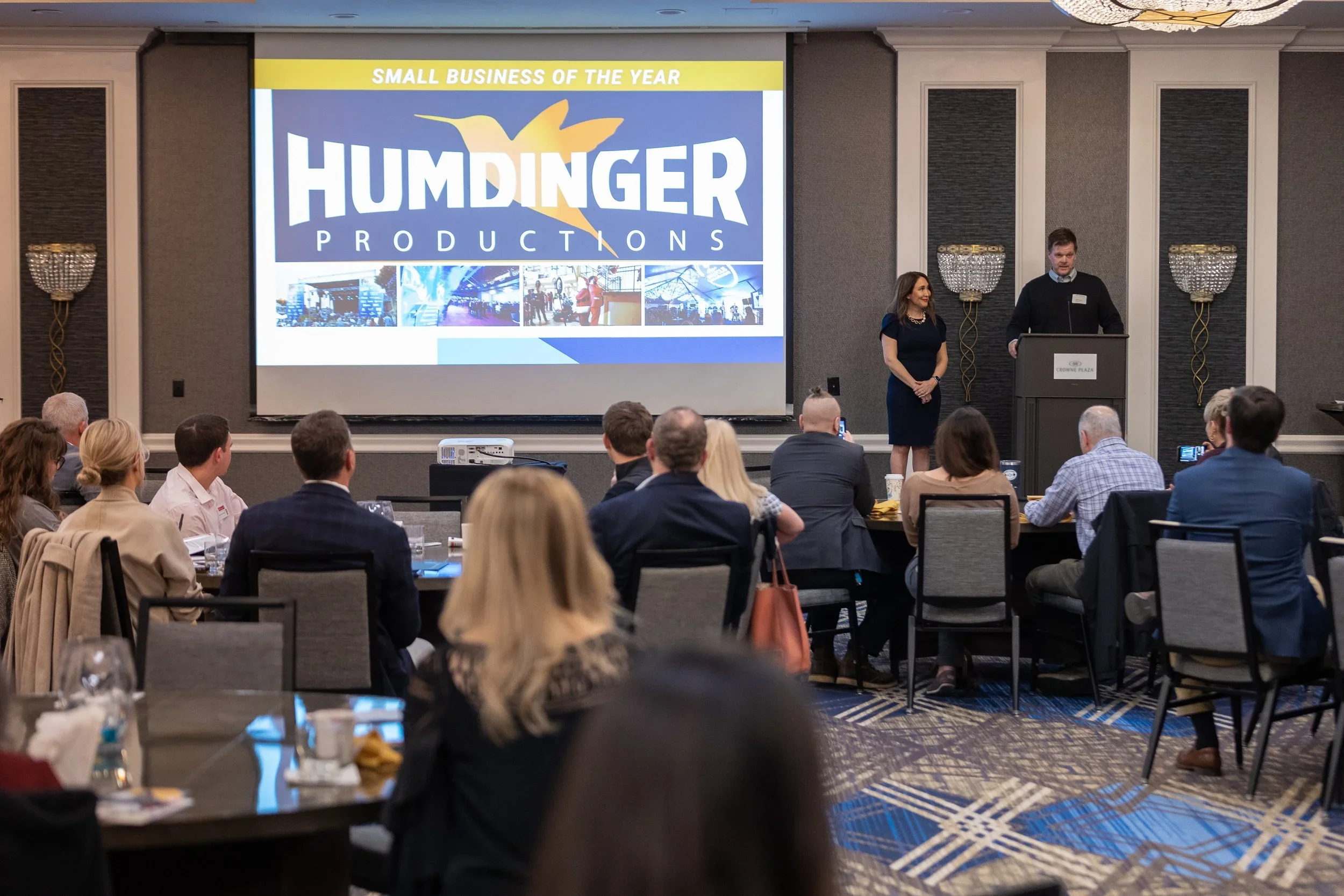 A conference room filled with attendees watching a presentation about Humdinger Productions, which was awarded Small Business of the Year. Two speakers stand at the front near a large screen displaying the company's logo and images, with seated audie