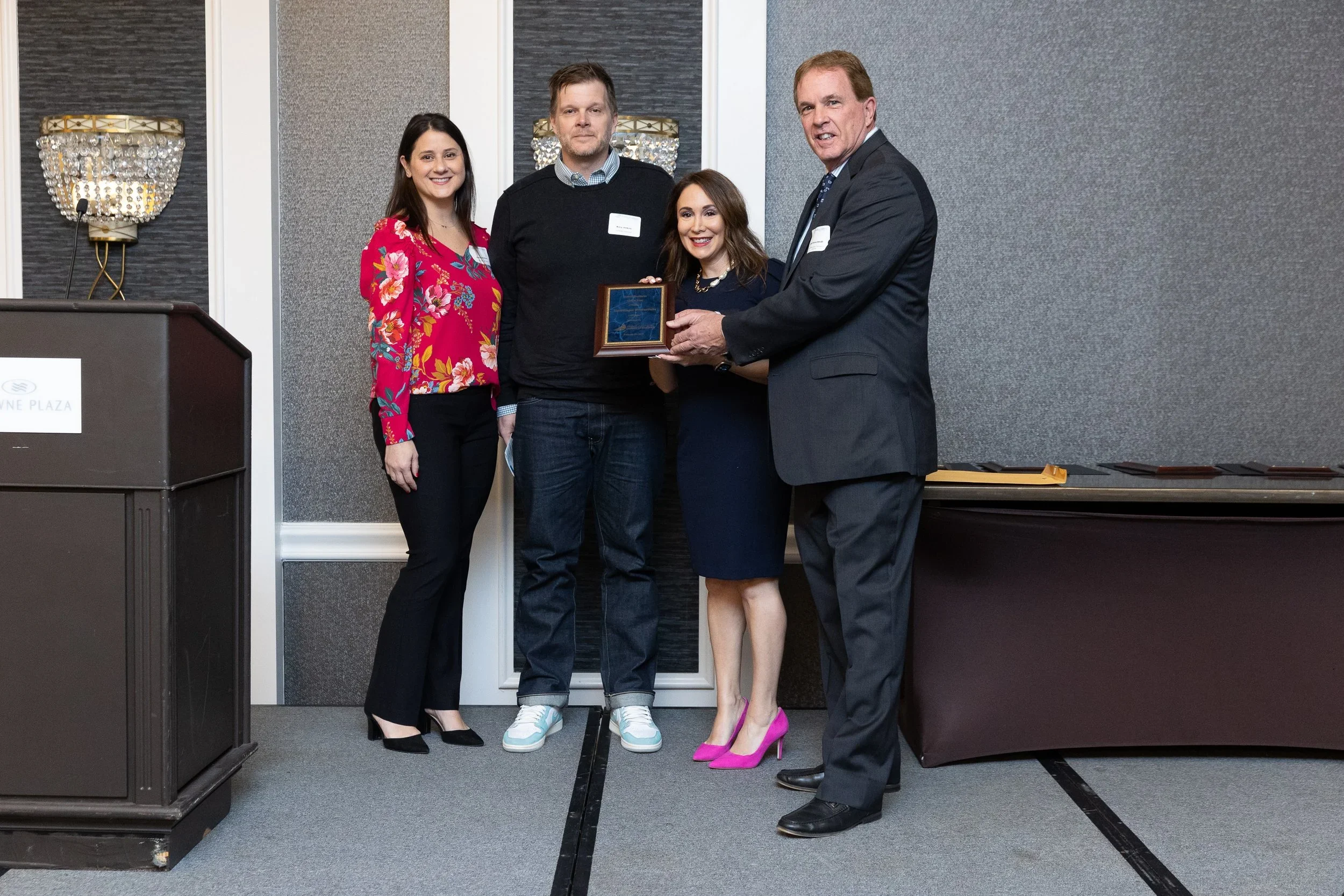 Five people standing together in a formal setting, with one woman holding a plaque and receiving an award from a man in a suit.