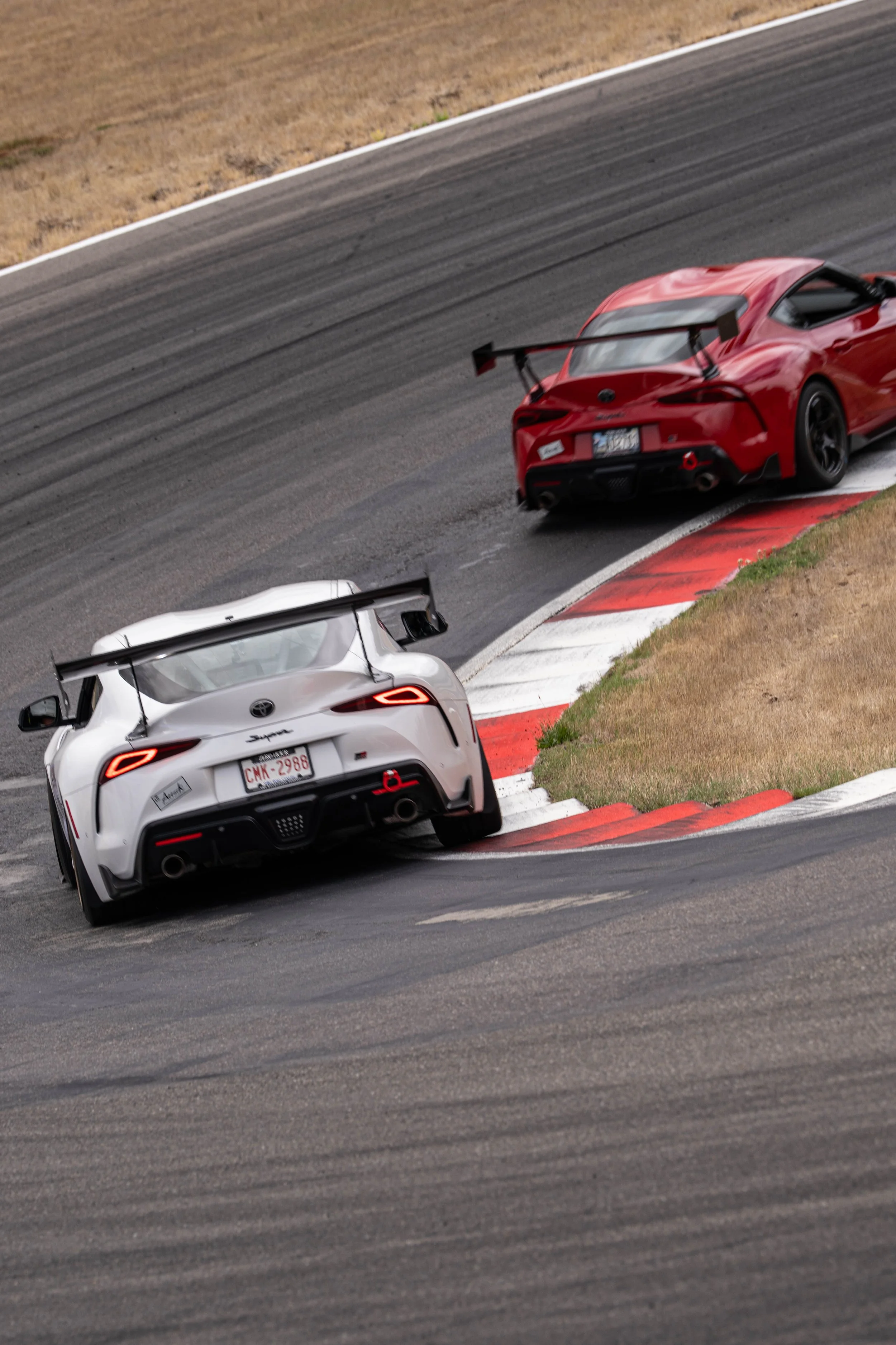 Two race cars, a white one and a red one, taking a sharp turn on a race track, with the red car near the curb and the white car following behind.