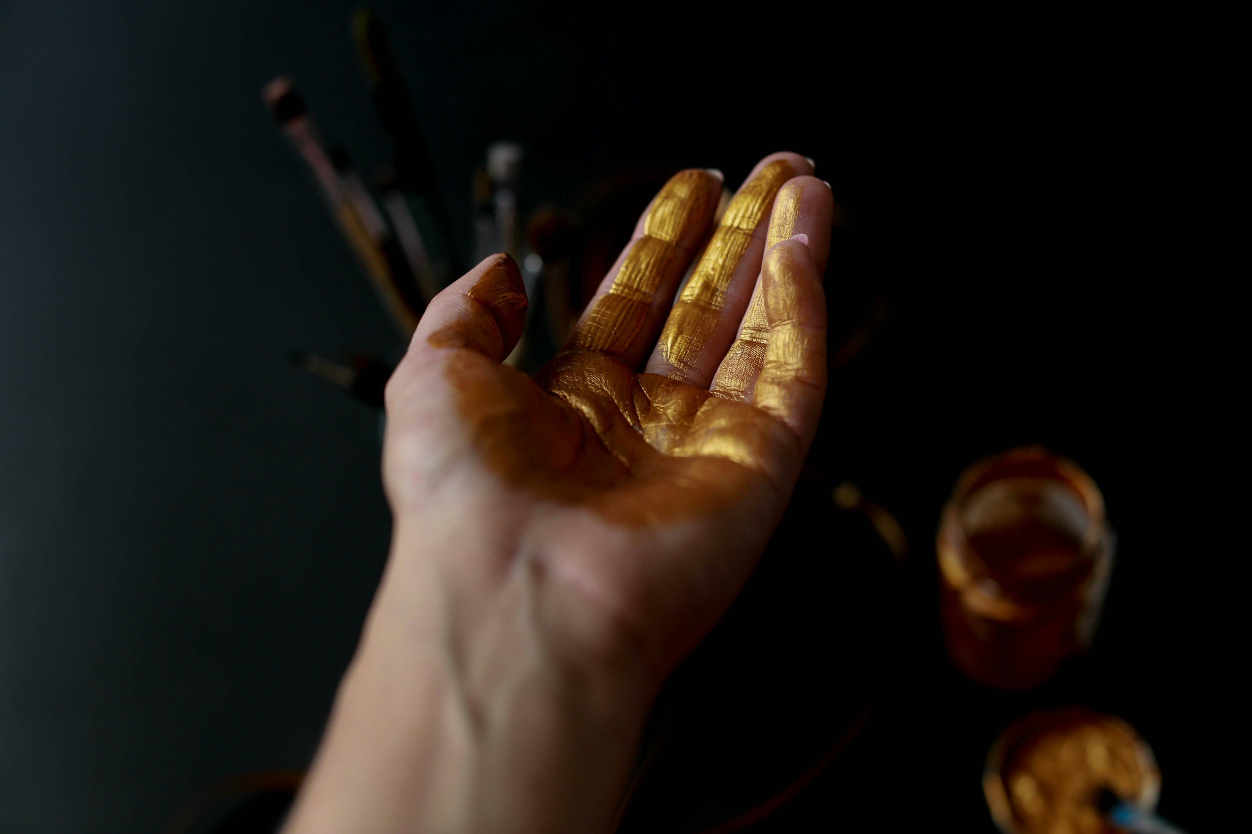 A hand is covered in gold paint, with paints and brushes in the background.