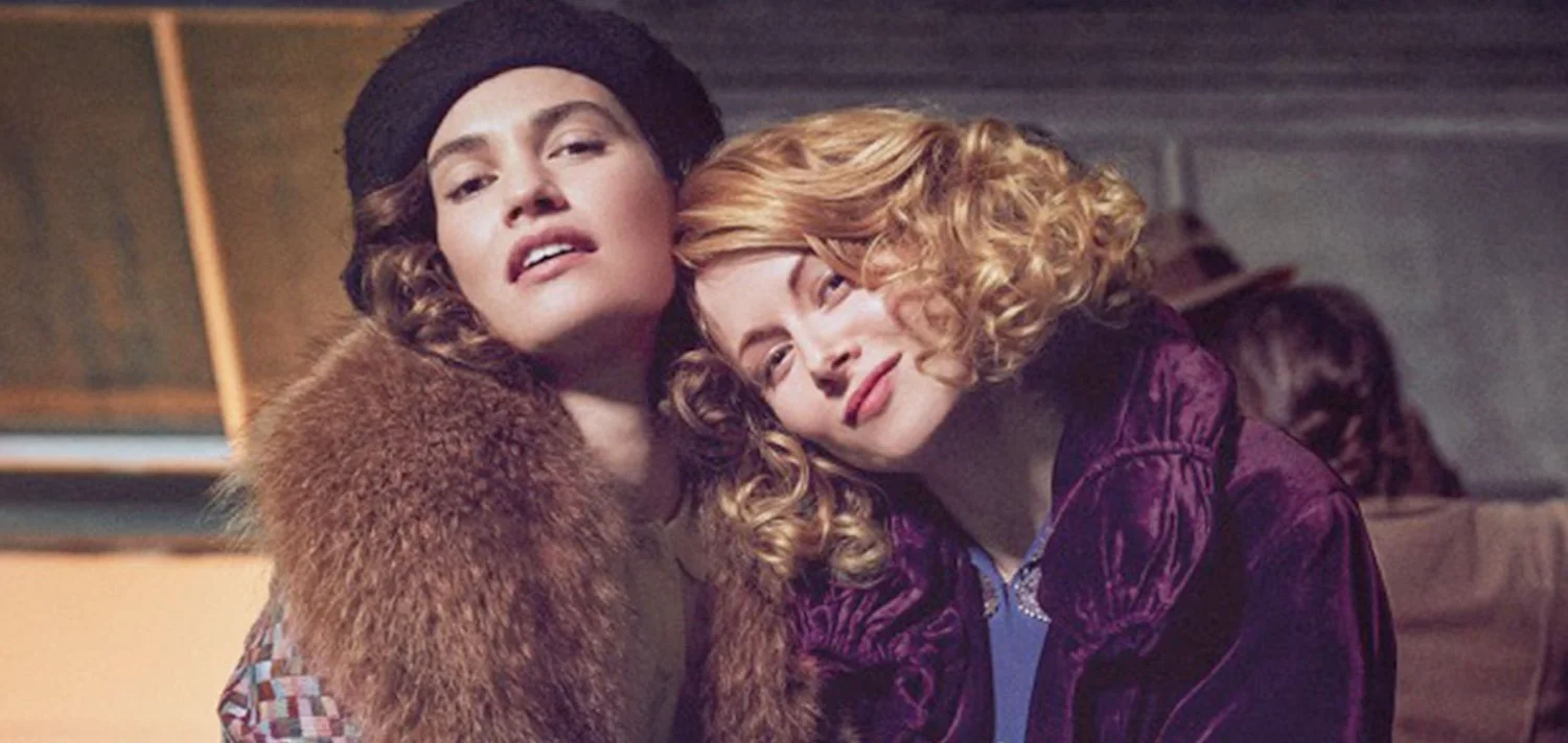 Two women with curly hair, one with dark cavan, the other with red hair, sitting closely together with affectionate expressions in a film by King Bee Productions. 