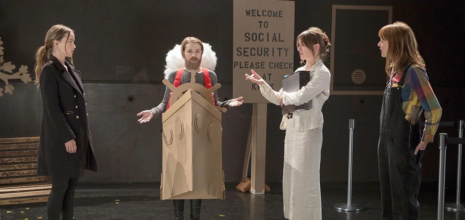 Dolly Wells and Emily Mortimer in a comedic airport security scene from the HBO series Doll & Em featuring a cardboard box costume with clouds and a ship’s wheel.