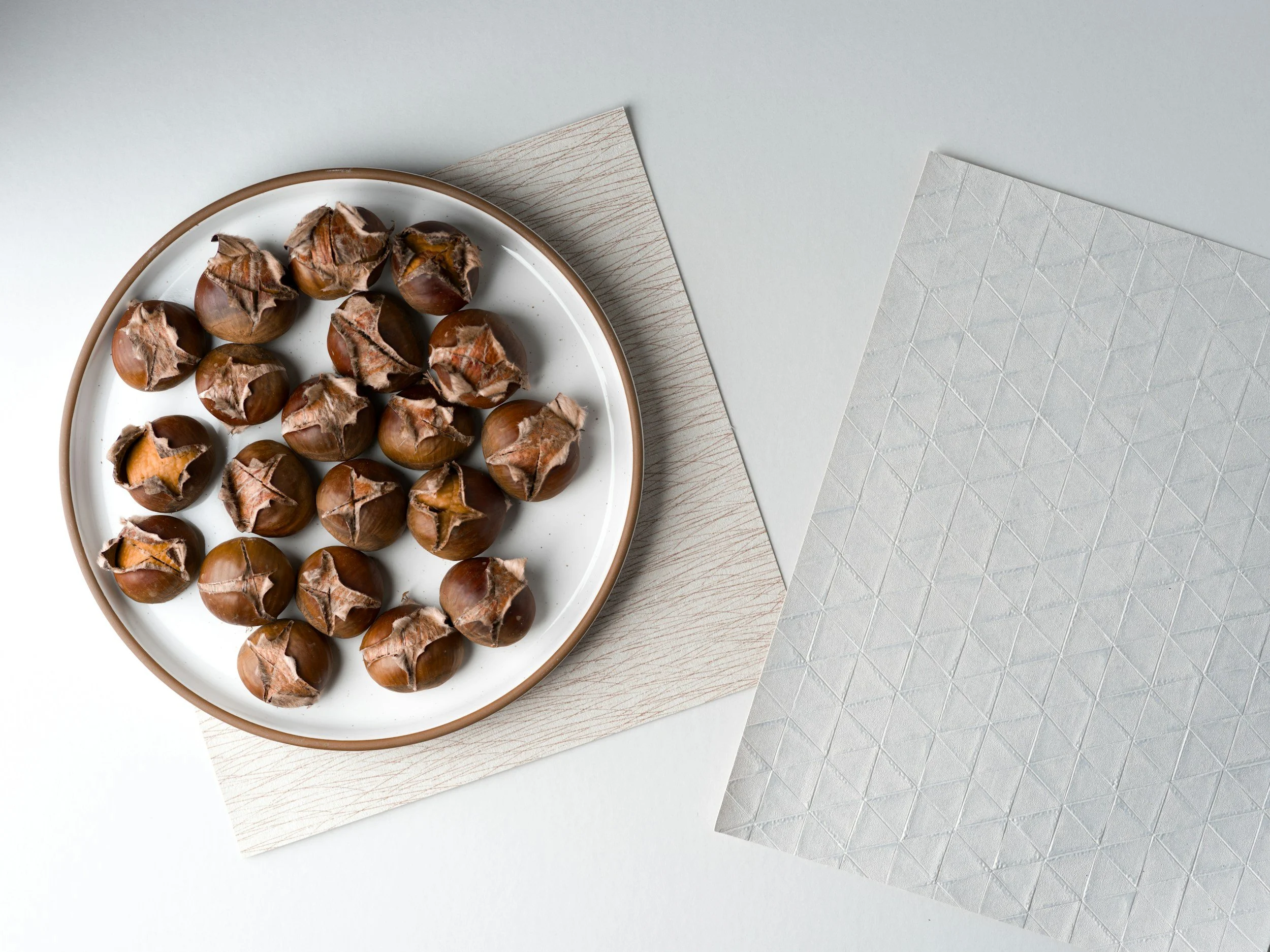 A white plate with roasted chestnuts on multi-colored textured paper, on a light background.