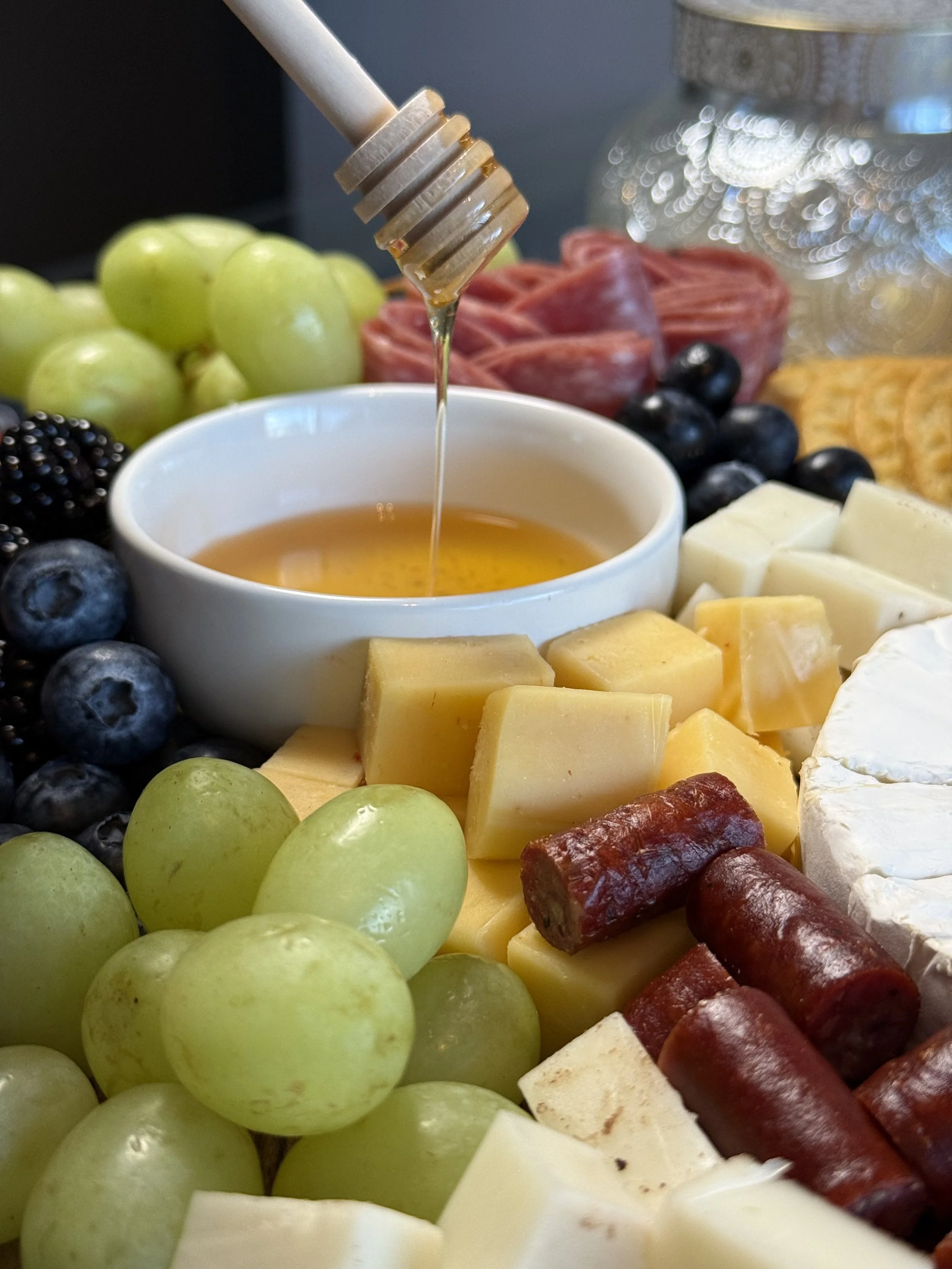 A cheese and fruit platter with honey, green grapes, blackberries, blueberries, sliced cheeses, and slices of cured meats.