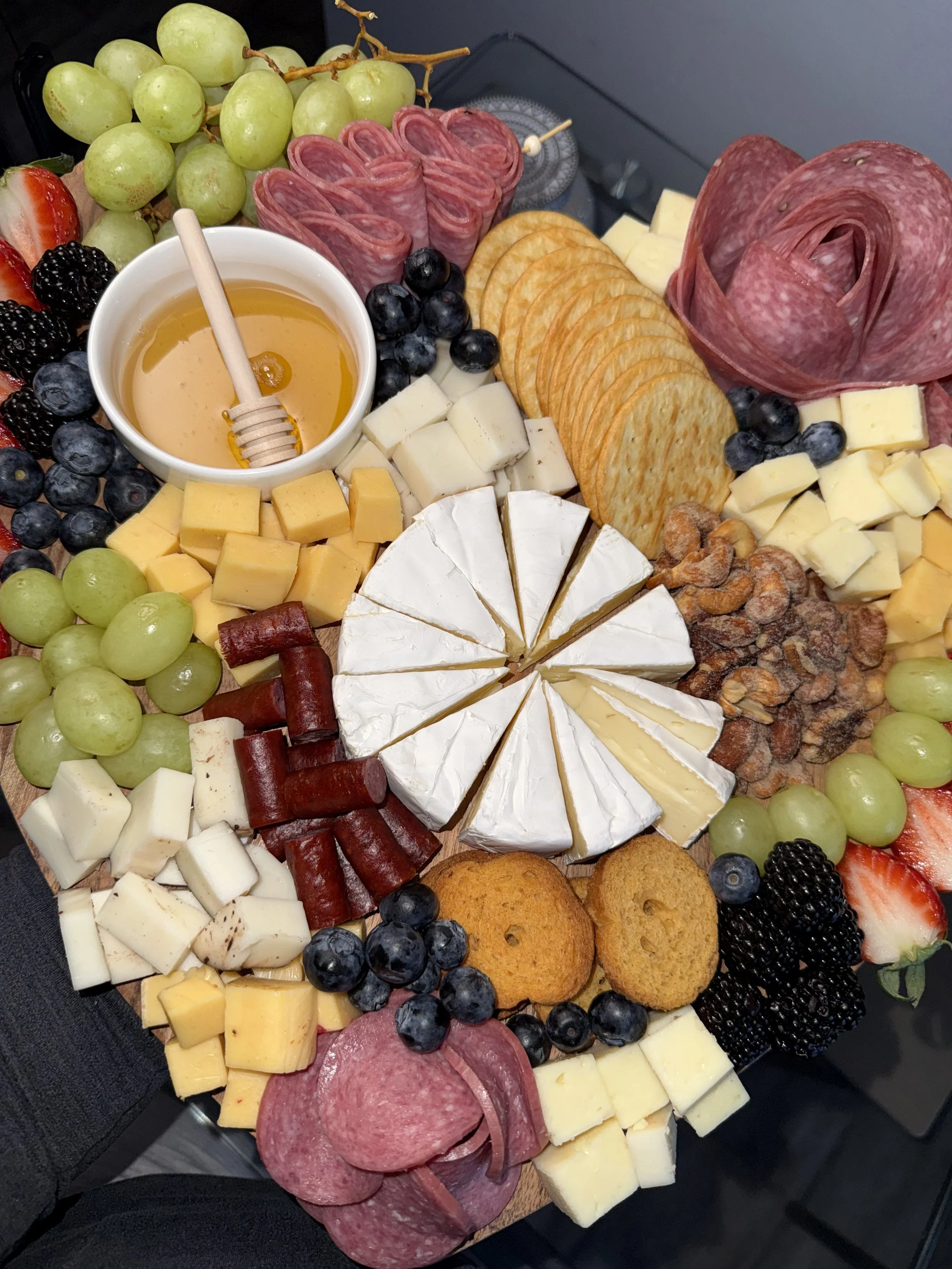 Charcuterie board with assorted cheeses, cured meats, crackers, honey, and fresh fruit including grapes, strawberries, blackberries, blueberries, and slices of melon and cheese.