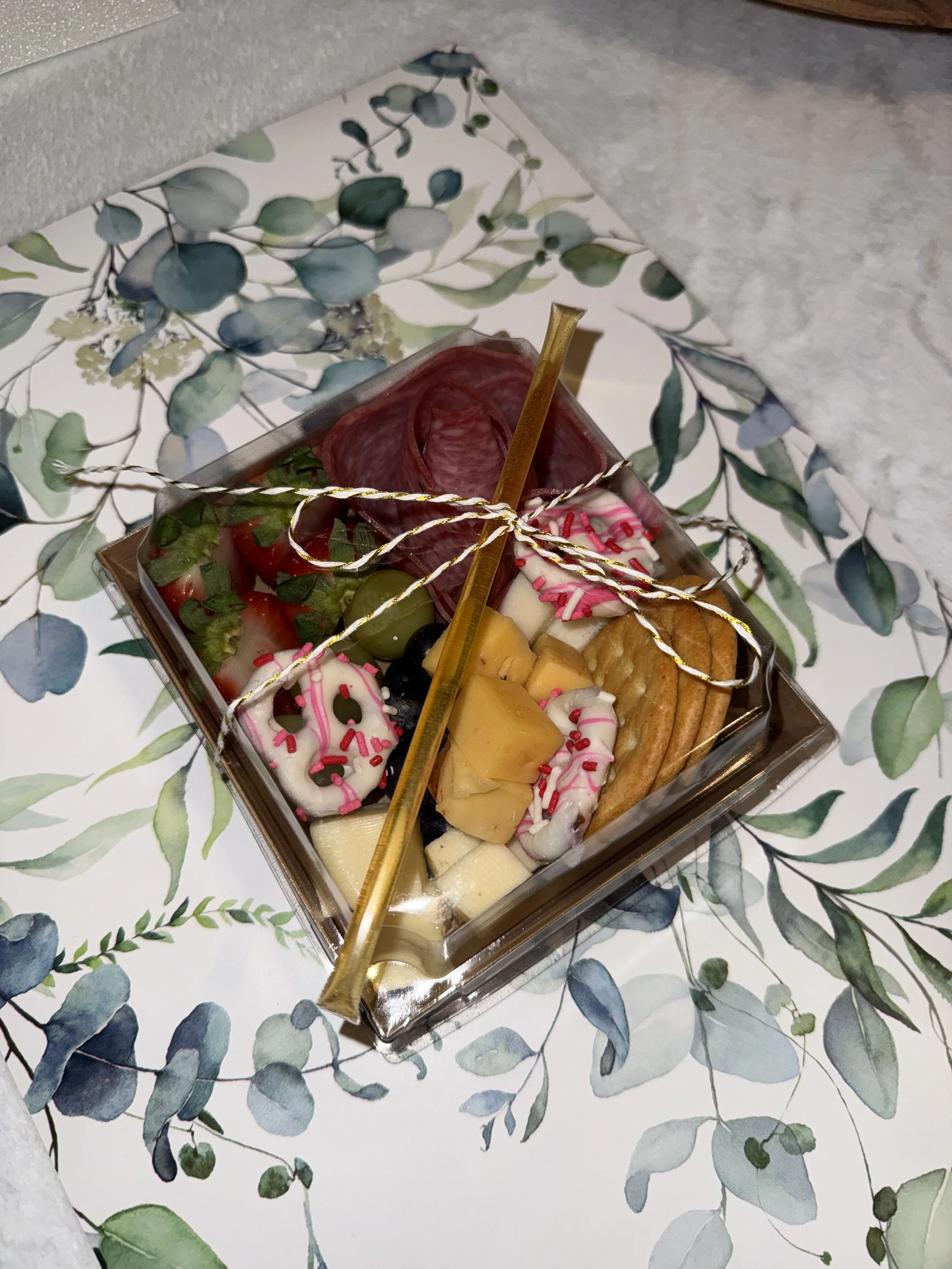 A clear plastic container filled with assorted cheeses, crackers, grapes, strawberries, salami, and decorated with pink and white sprinkles, topped with a gold spoon and gold and white ribbon, sitting on a decorative leaf-patterned paper.