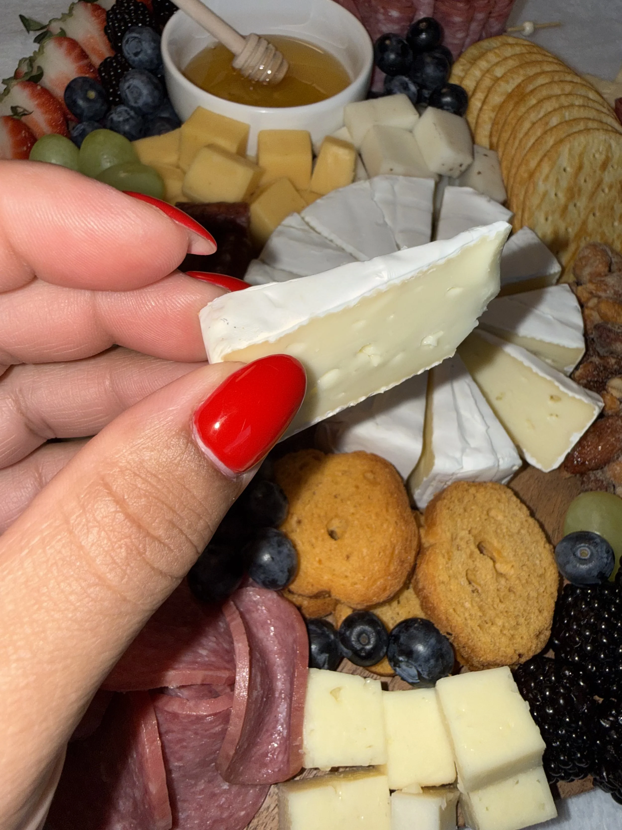 Close-up of a hand with red painted nails holding a wedge of white brie cheese with a variety of cheese, bread, grapes, blackberries, blueberries, strawberries, honey, and cured meats on a charcuterie board.