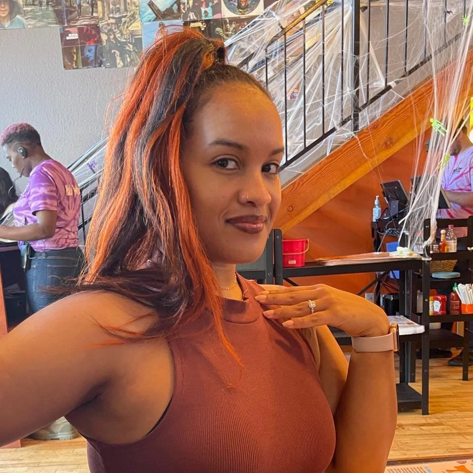 Ashleigh Sanders, founder of Bites of Bliss charcuterie, a woman with long brown hair in a ponytail, wearing a sleeveless brown top, posing indoors with one hand on her shoulder.