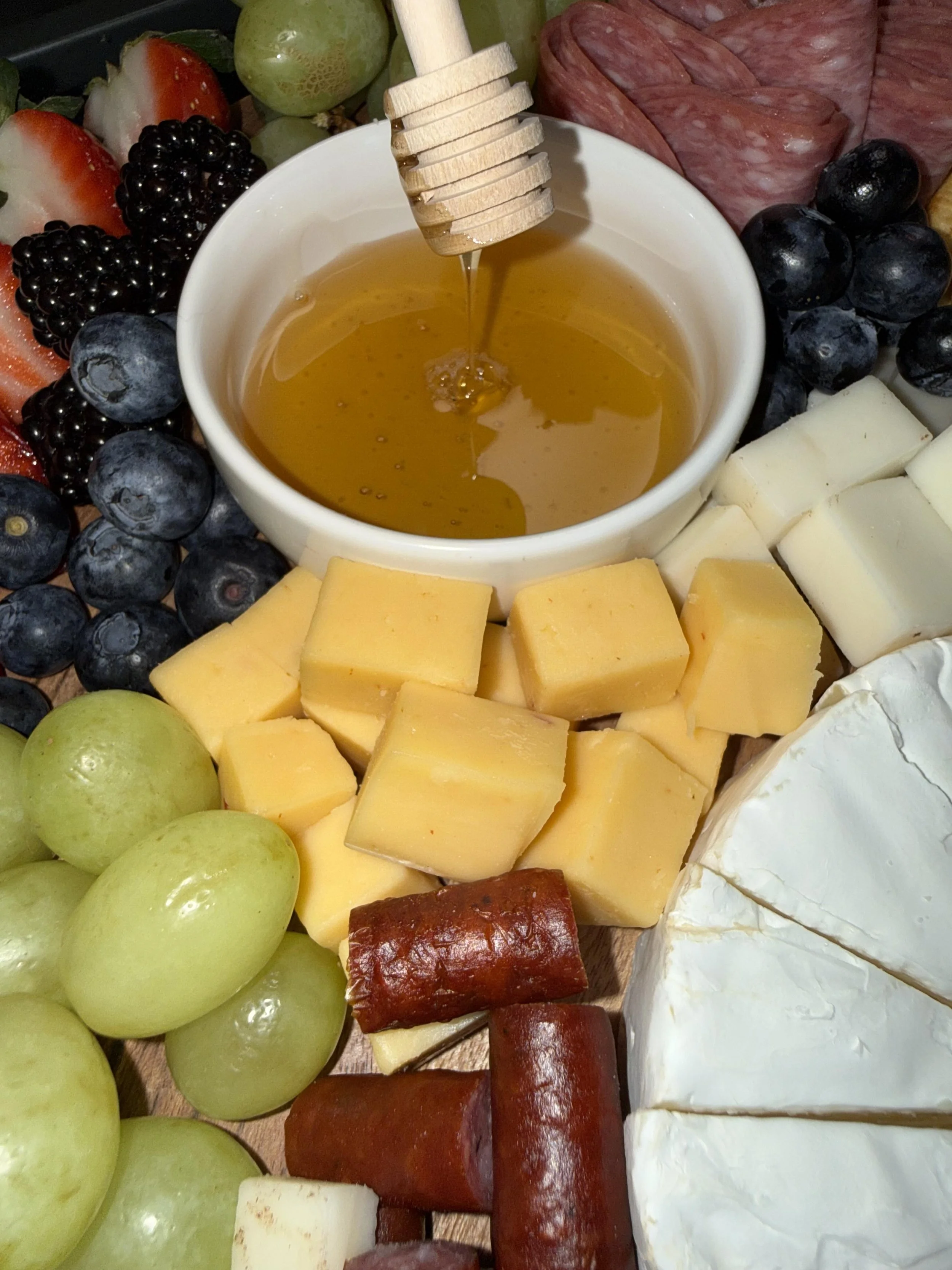 A cheese and fruit platter with honey. The platter includes green grapes, blueberries, blackberries, sliced strawberries, sliced cheese, cubed yellow cheese, sliced salami, and a wheel of brie cheese. Honey is drizzling from a honey dipper into a whi